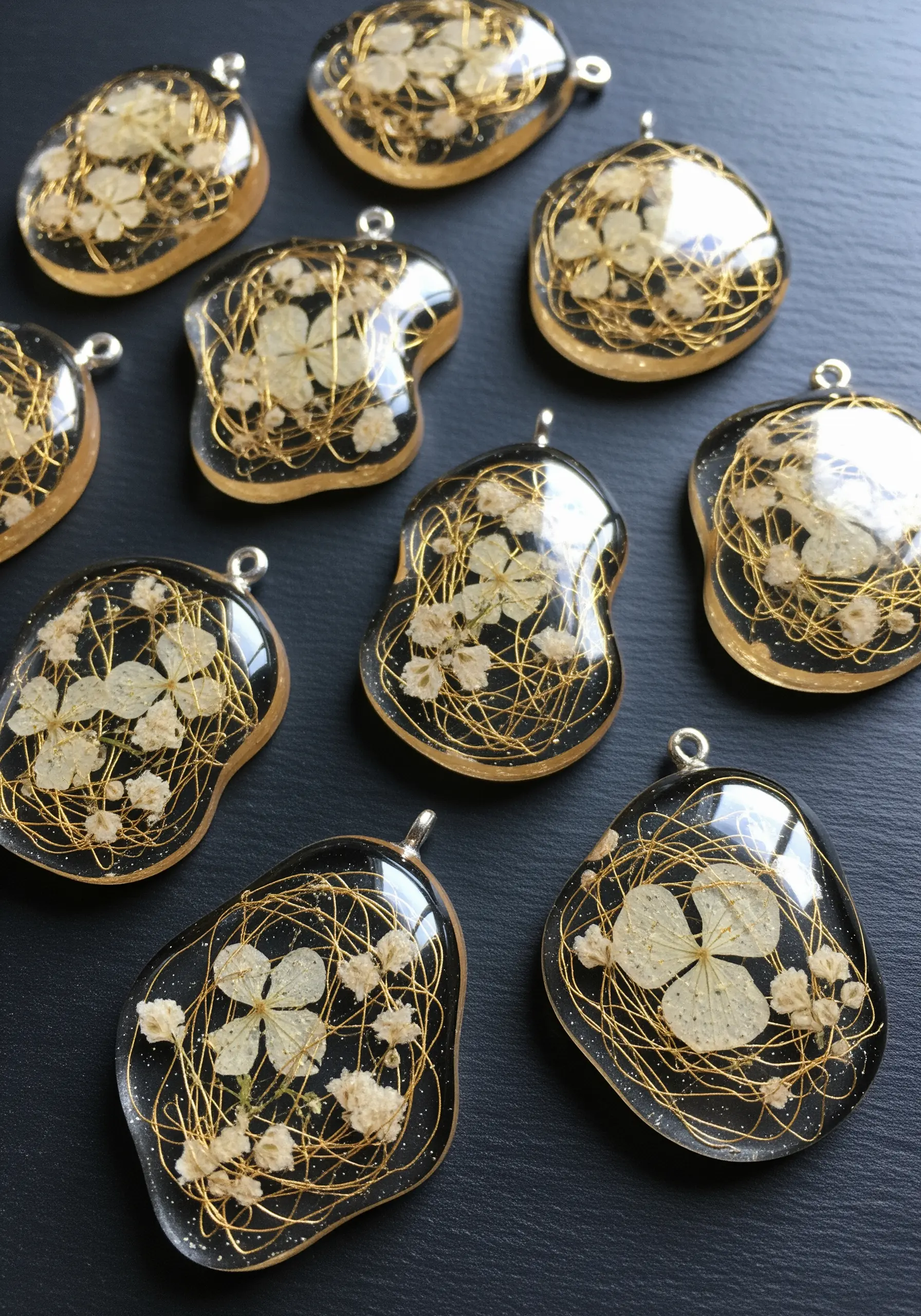 Resin pendants with dried white flowers and tangled gold thread on a black background.