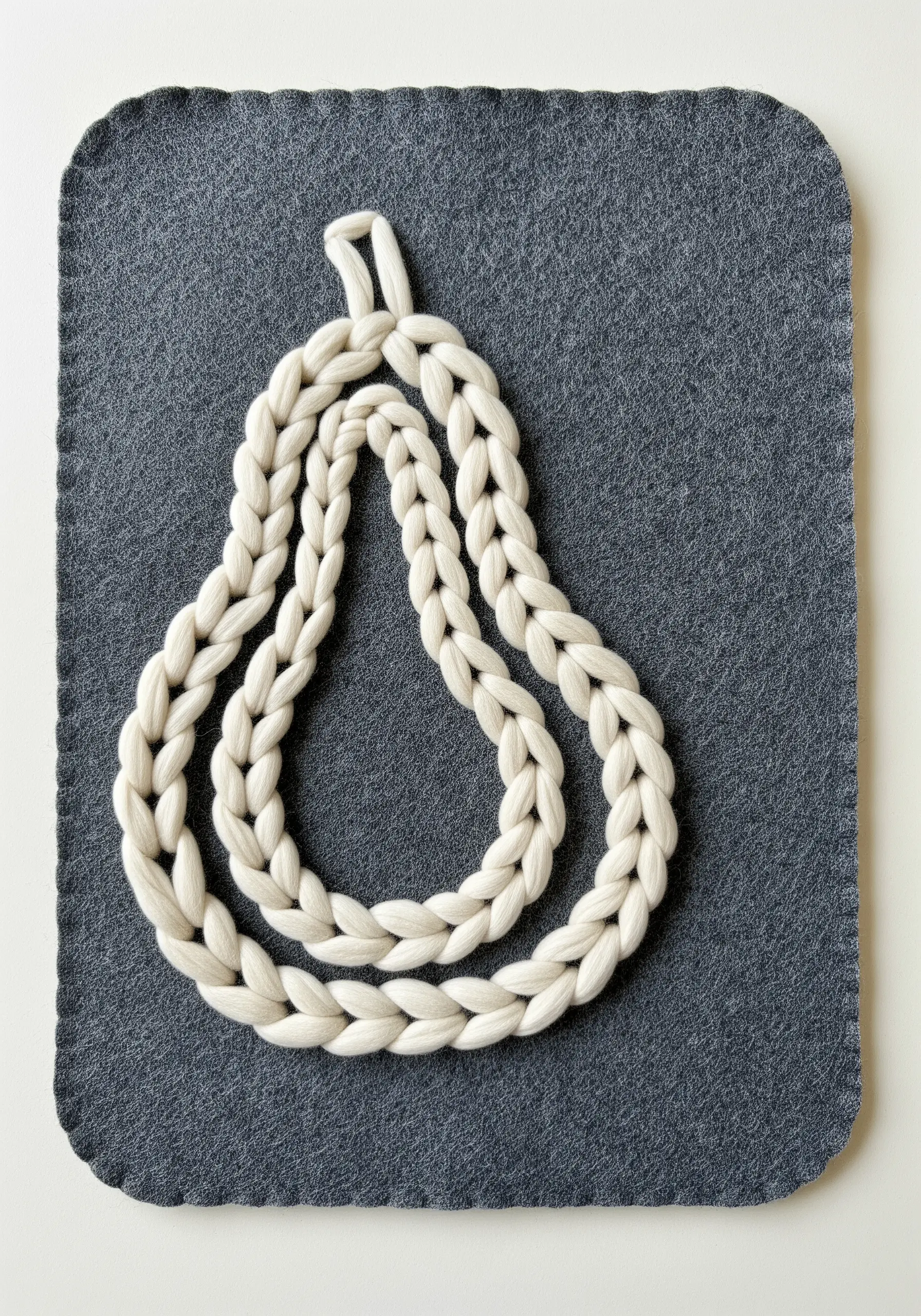 A thick, braided off-white yarn couched in the shape of a pear on a dark grey felt placemat.