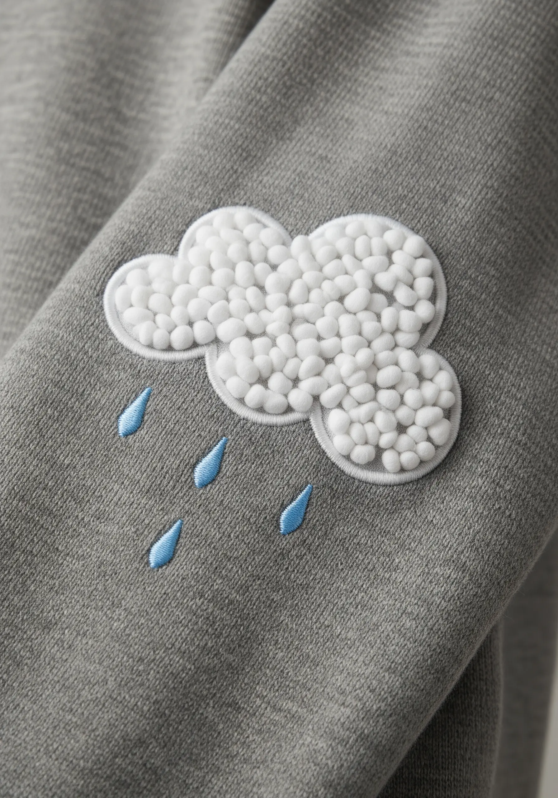 A fluffy cloud embroidered with white French knots and blue satin stitch raindrops on a grey sleeve.