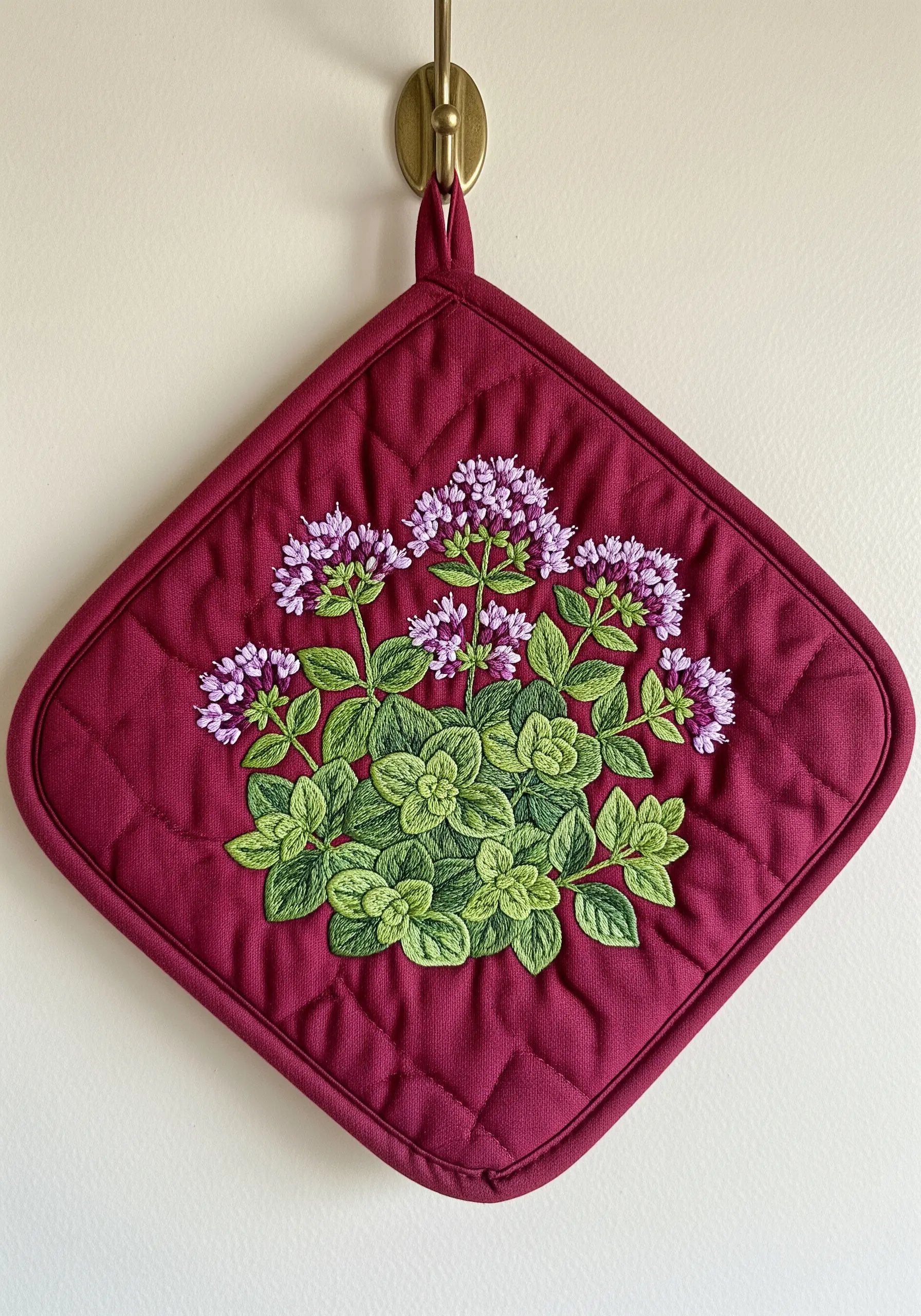 A dense and textured embroidery of flowering oregano on a quilted burgundy potholder.