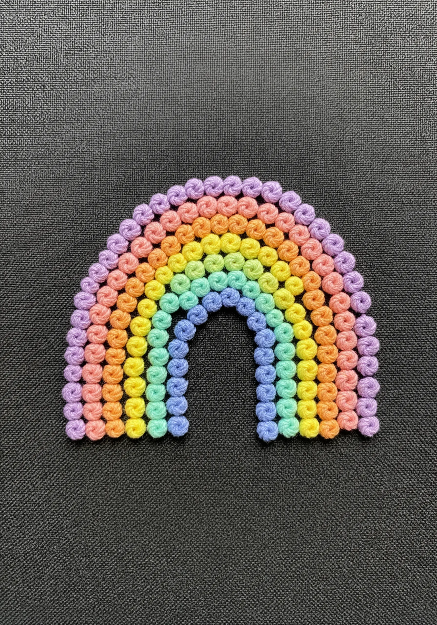 A pastel rainbow made entirely of tightly packed, colorful bullion knots on black fabric.