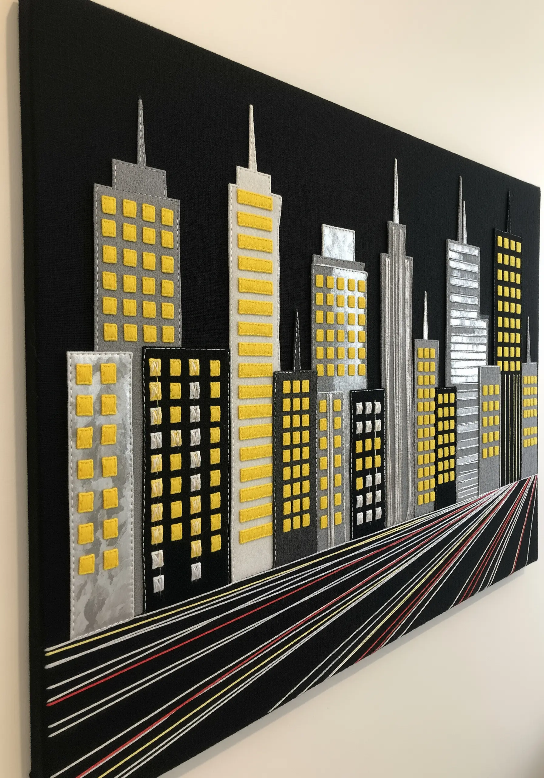 A modern cityscape wall art made with fabric appliqué buildings and long stitched light trails.