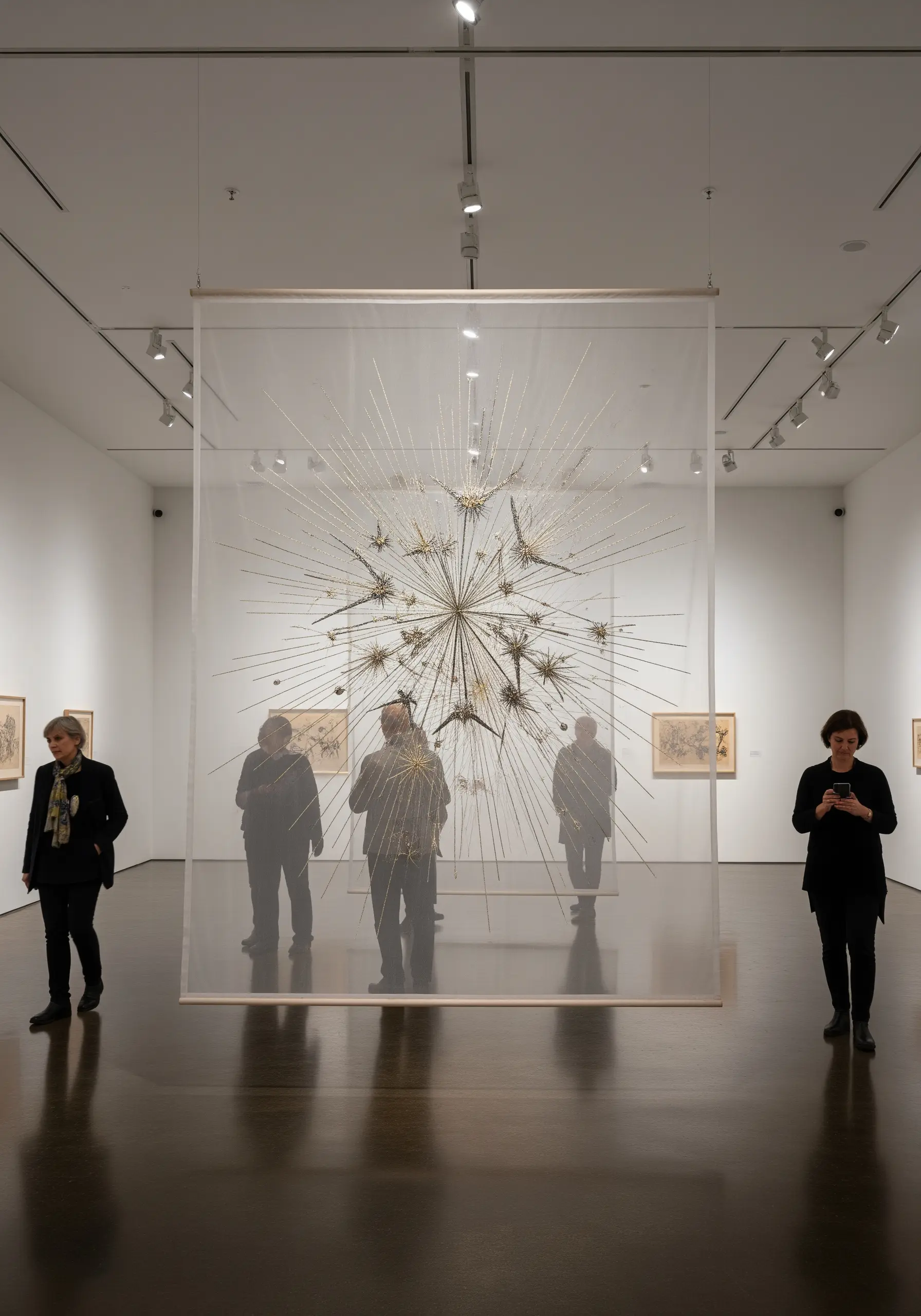 A suspended starburst sculpture in a glass case, made of radiating rods and metallic thread.