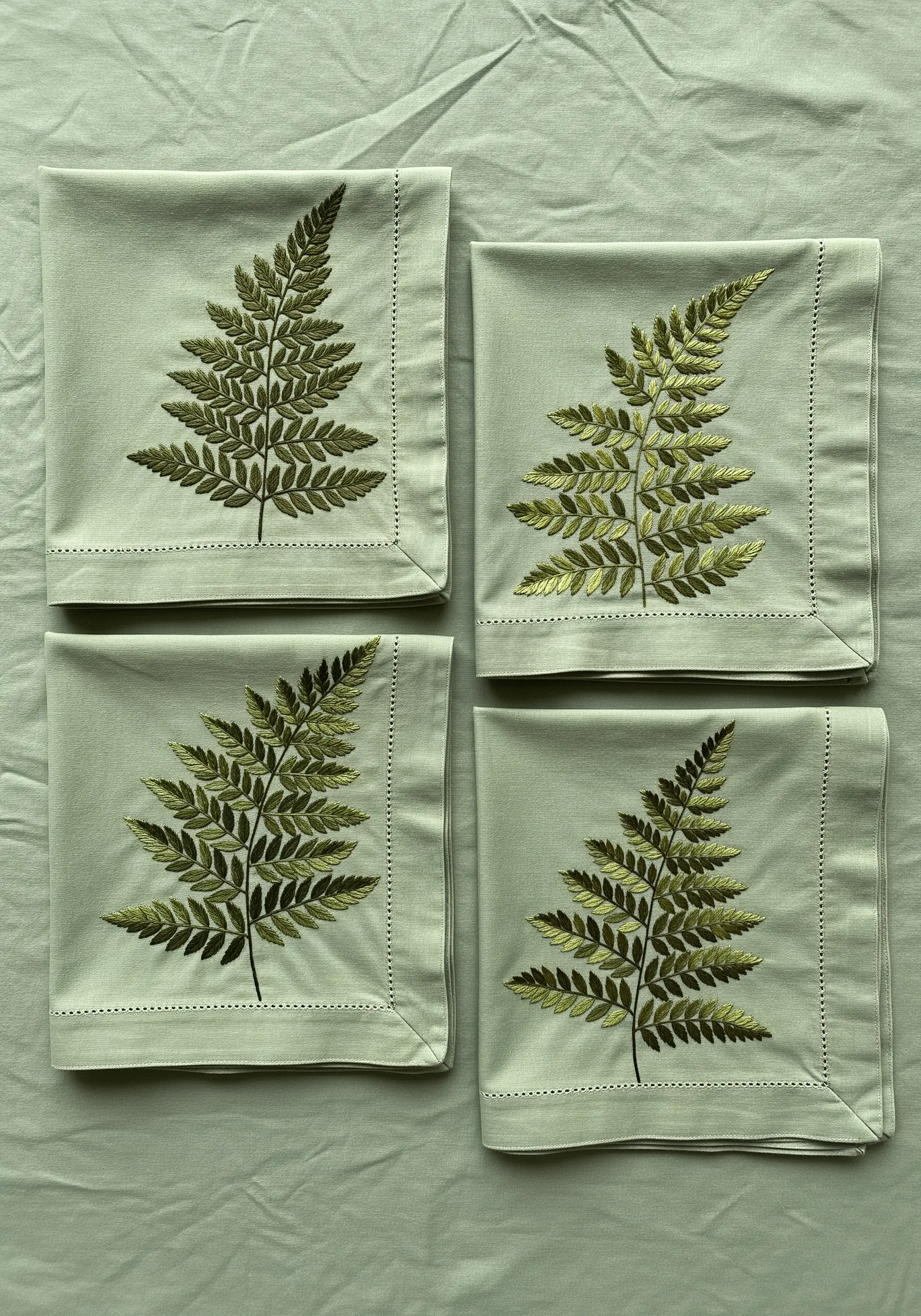Four sage green napkins, each embroidered with a realistic, multi-toned fern leaf.