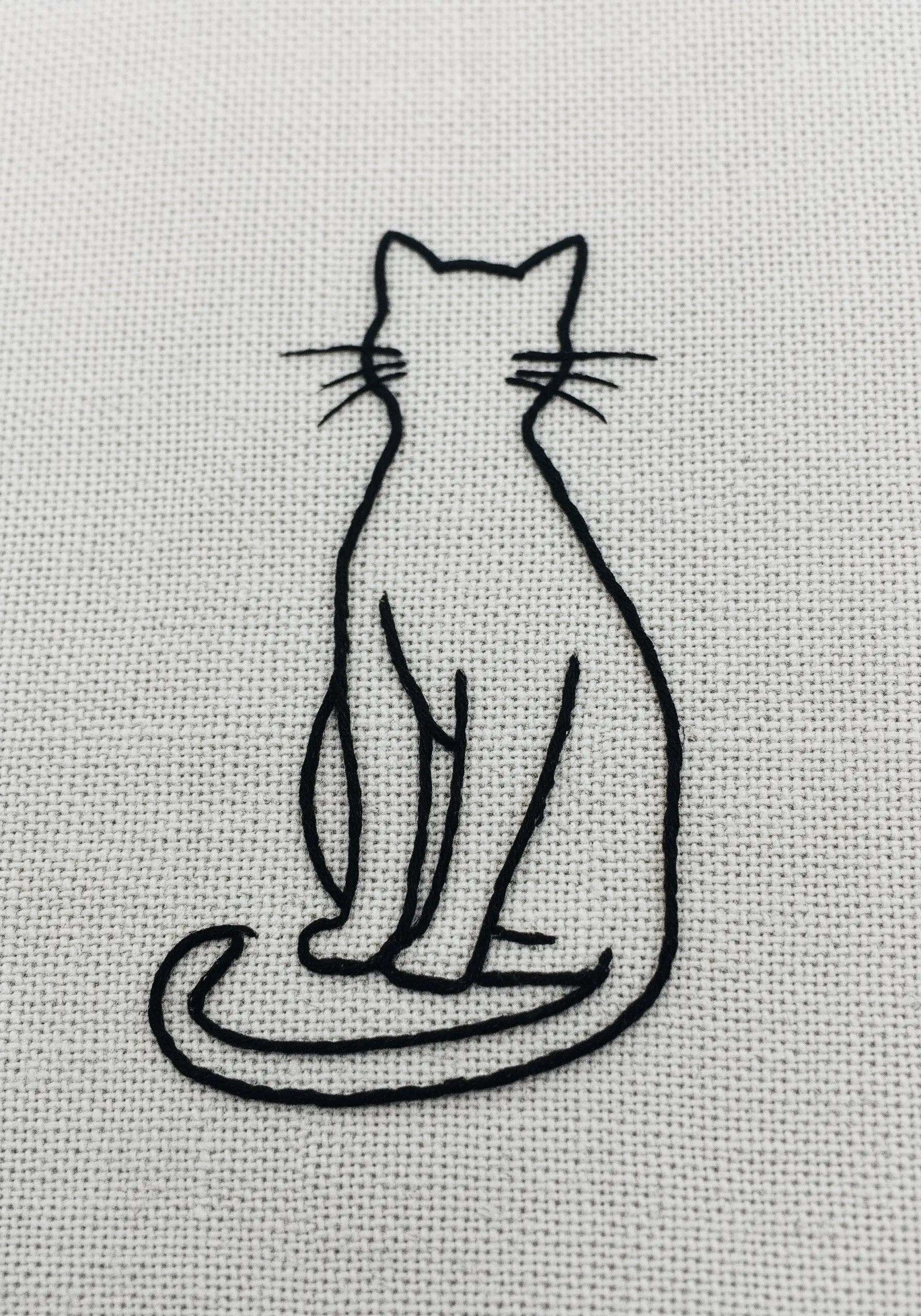 A minimalist black outline of a sitting cat stitched on a light neutral fabric.
