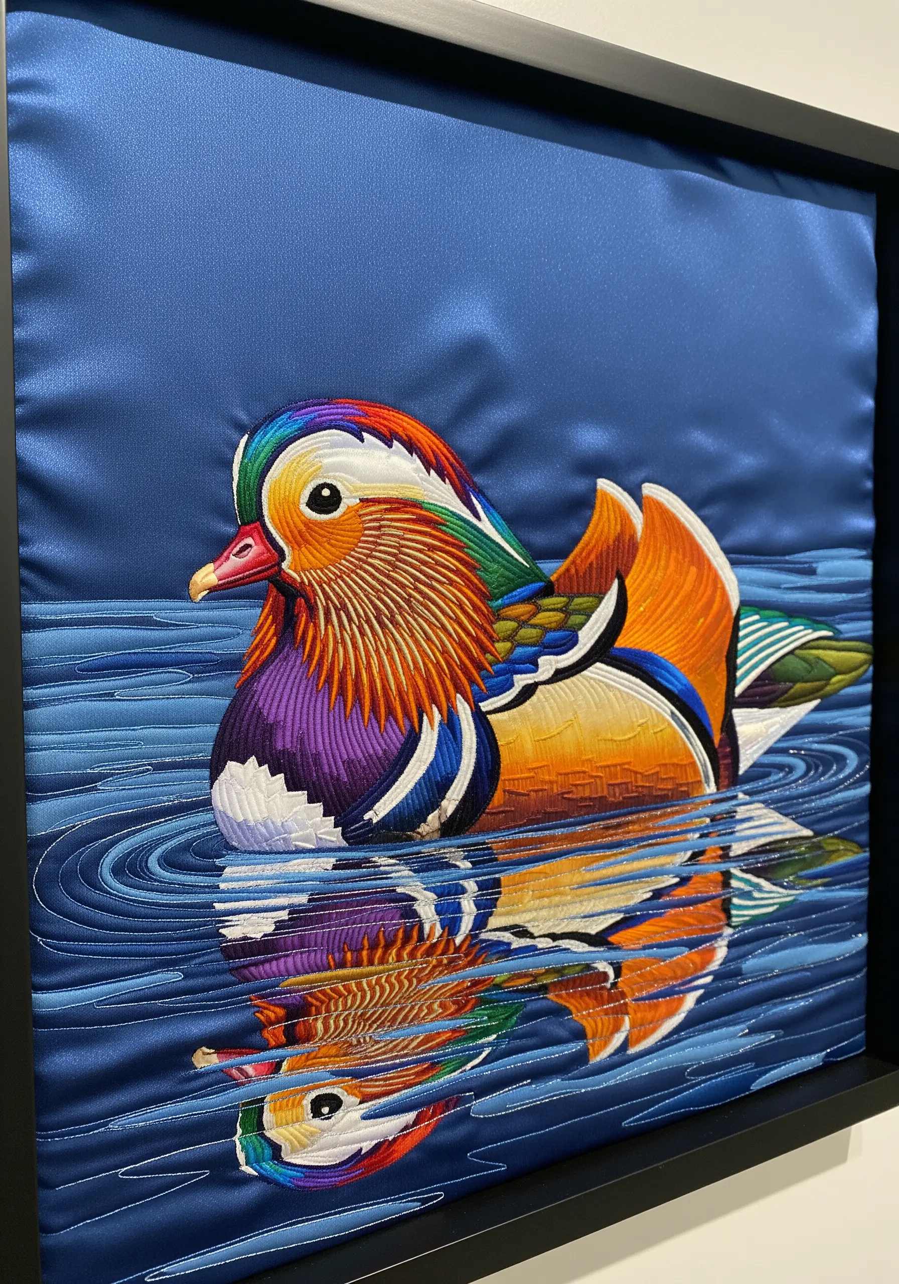 A vibrant mandarin duck thread-painted on blue satin fabric, with a stitched water reflection.