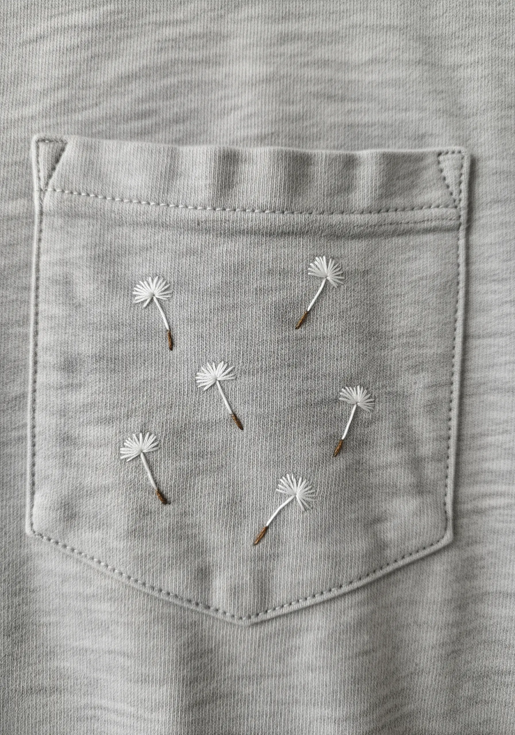 Delicate dandelion seeds made with detached chain stitches on a gray t-shirt pocket.