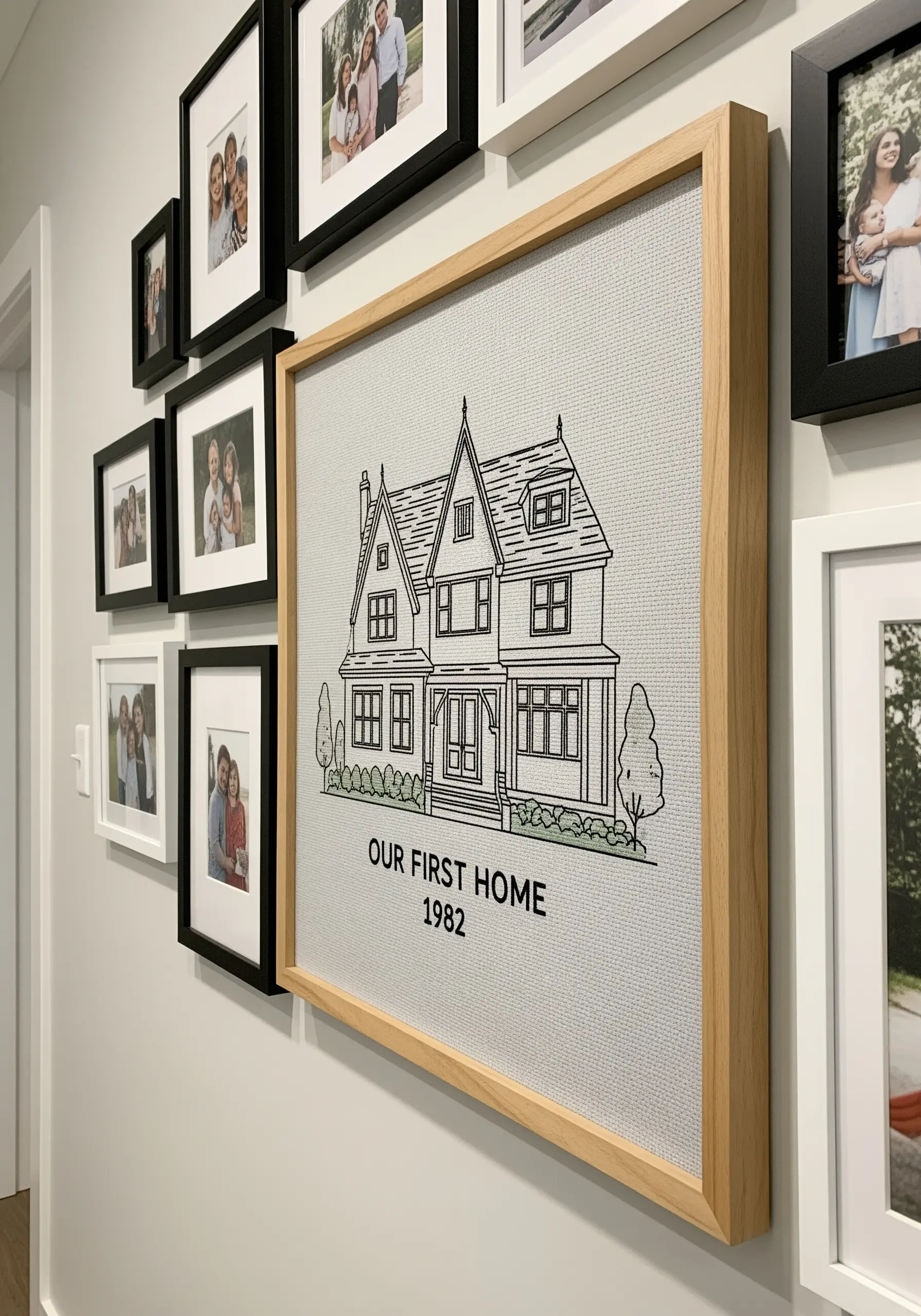 Framed line art embroidery of a house, hanging on a gallery wall with photos.