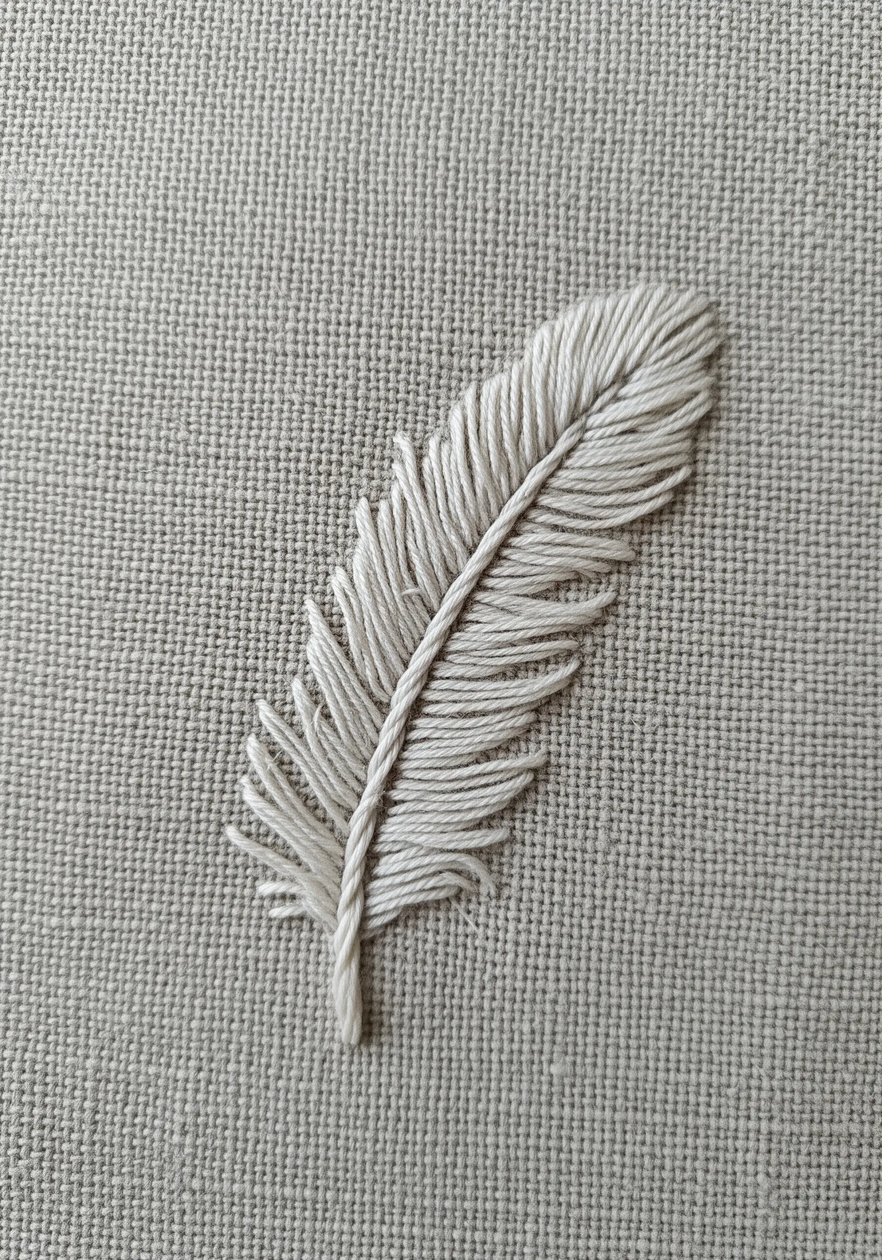 A single white feather embroidered on grey linen using the fishbone stitch technique.