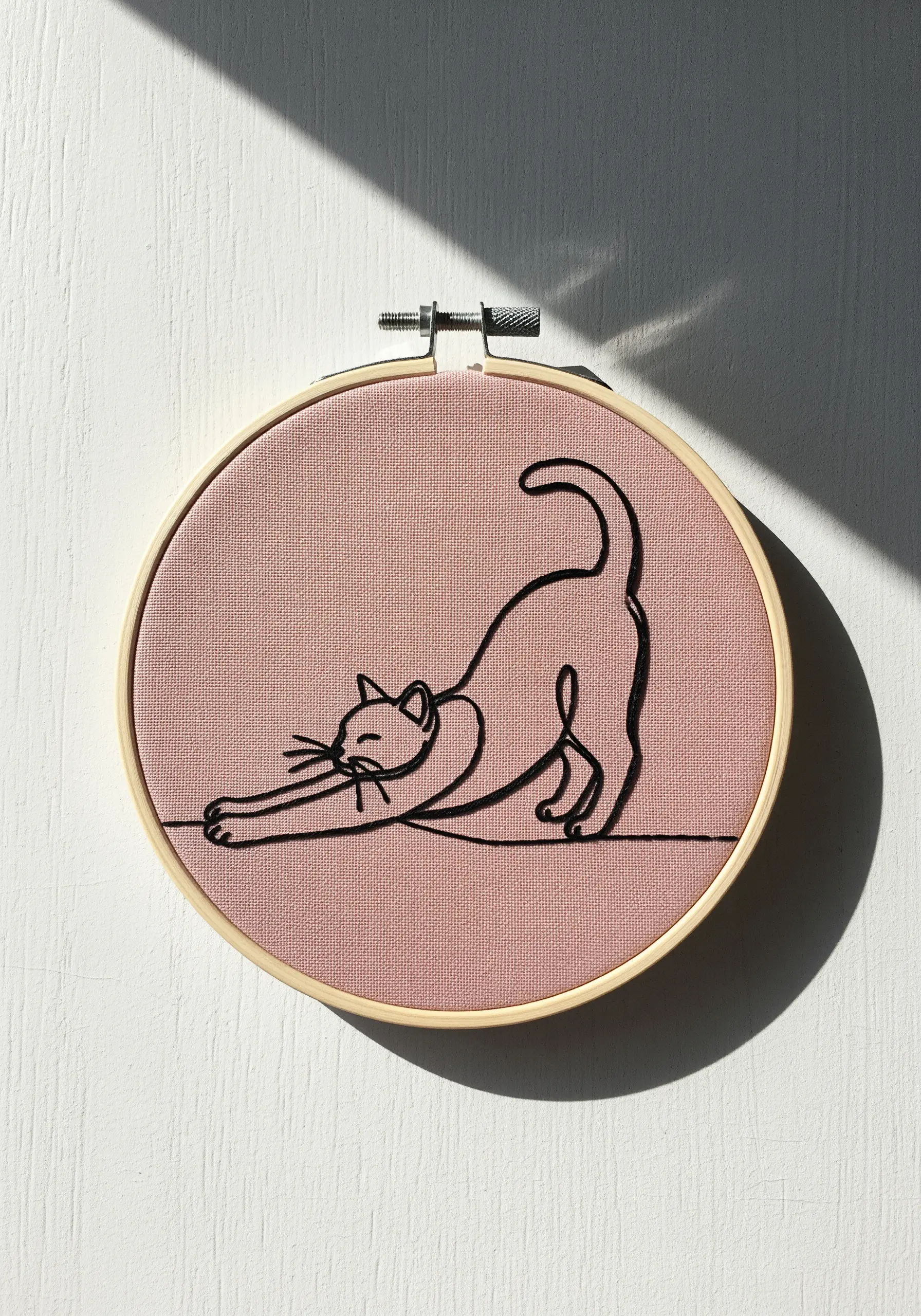 Minimalist line art embroidery of a cat stretching on a dusty pink background.
