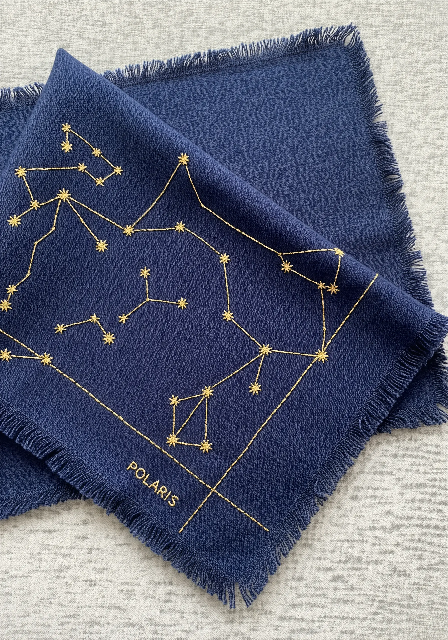 A gold-thread constellation map embroidered on a frayed-edge navy blue linen napkin.