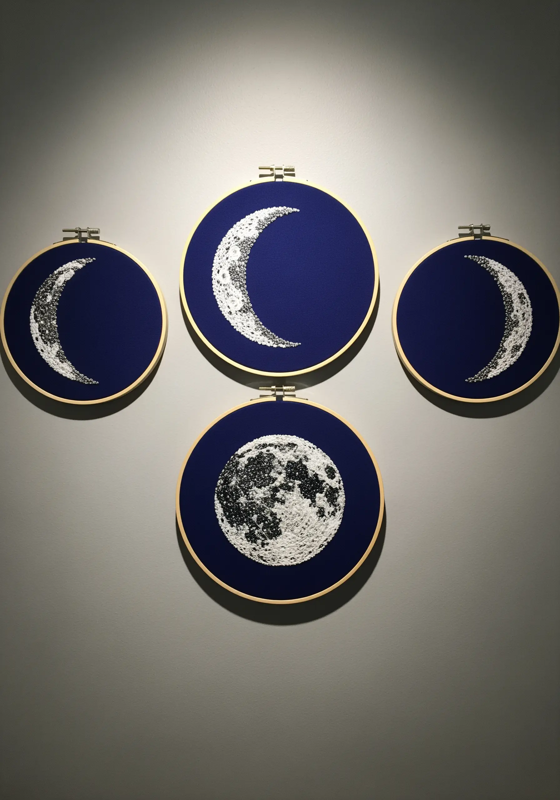 Four embroidery hoops showing moon phases created with white and grey French knots on navy fabric.