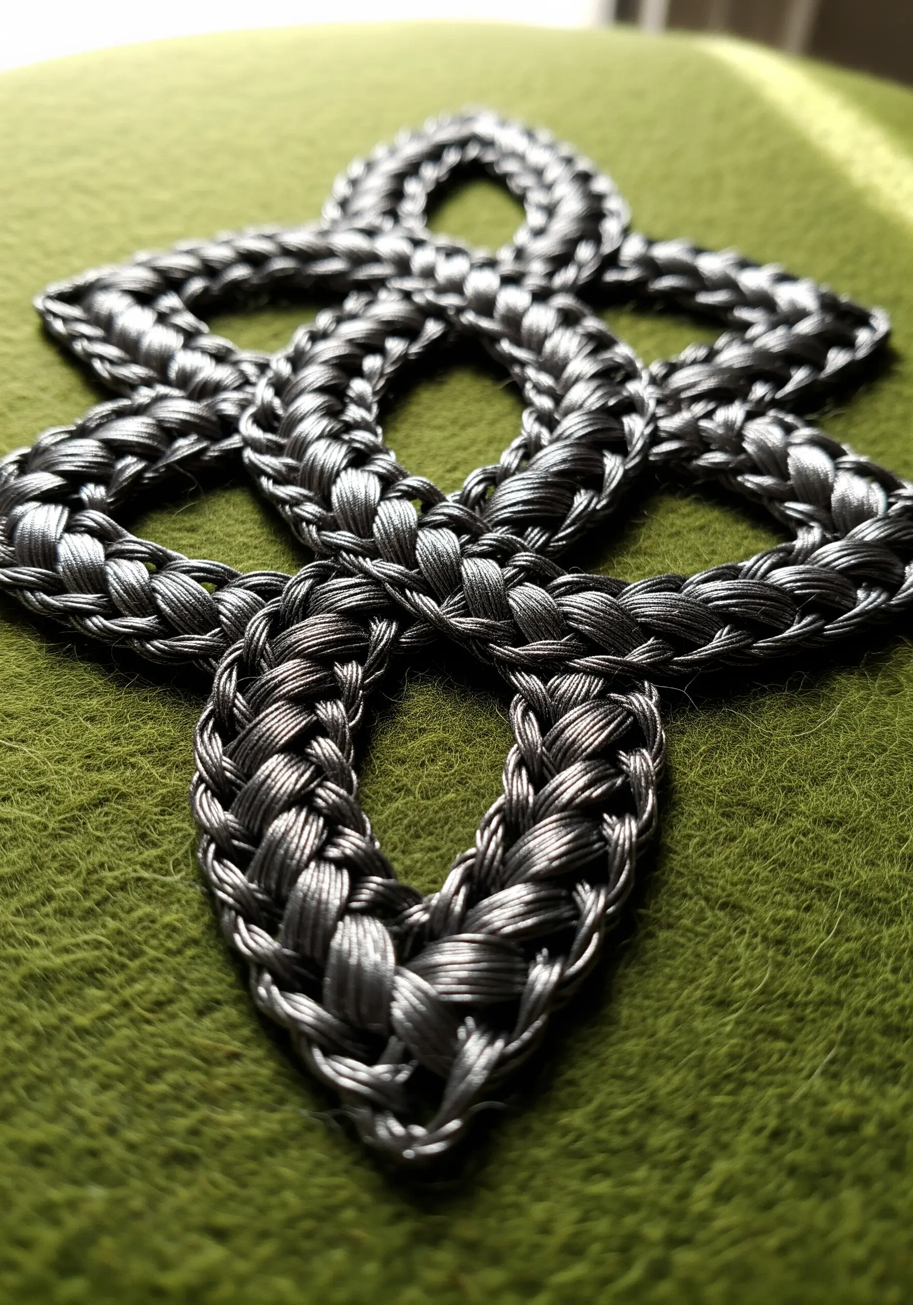 Close-up of a 3D Celtic knot embroidered with thick, metallic silver thread on green felt.