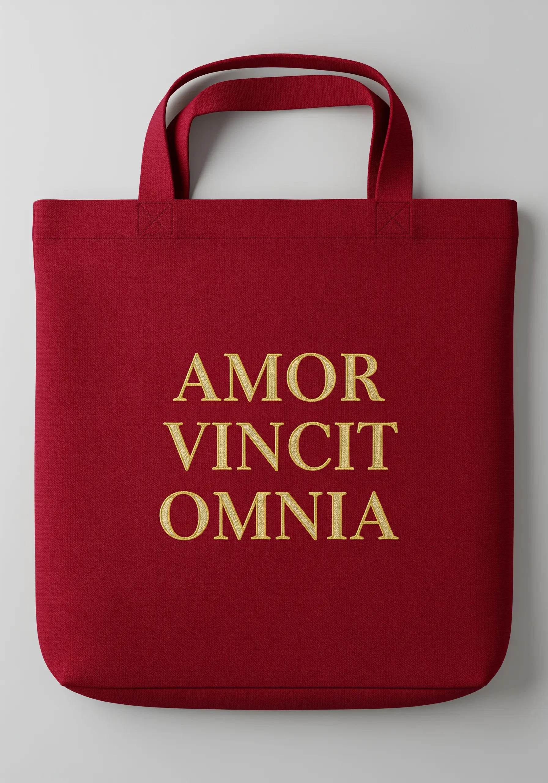 The Latin phrase 'Amor Vincit Omnia' embroidered in gold serif font on a deep red tote bag.