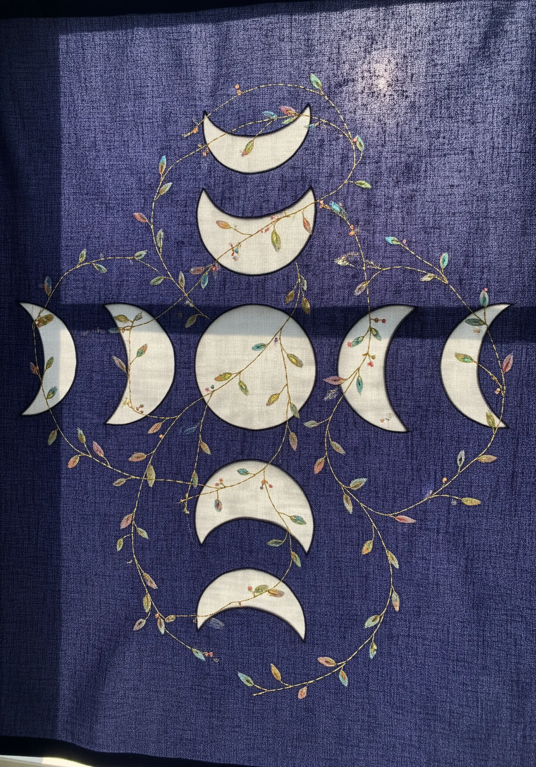 A moon phase design on blue fabric, combining white appliqué moons and embroidered vines.