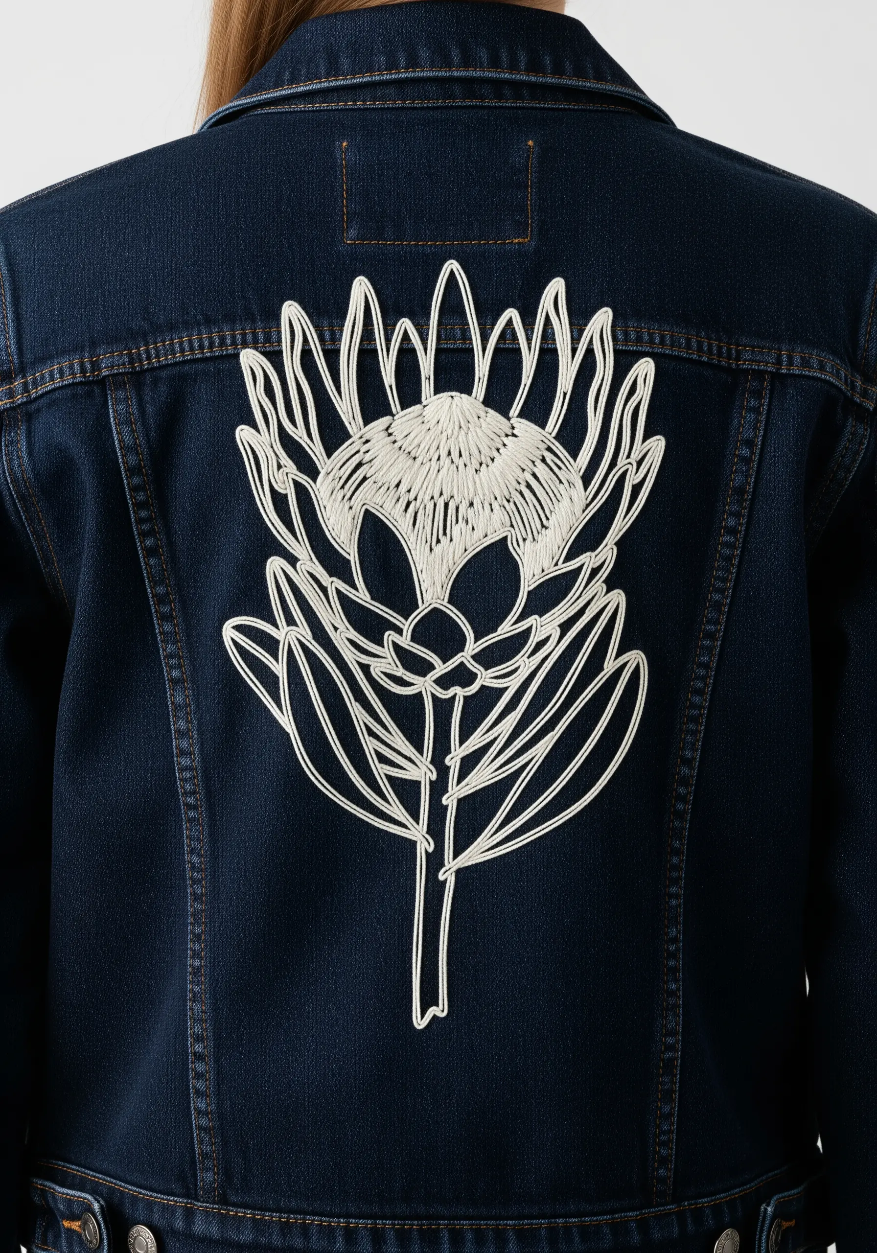 A large protea flower embroidered in white line art and fill stitches on a dark denim jacket.