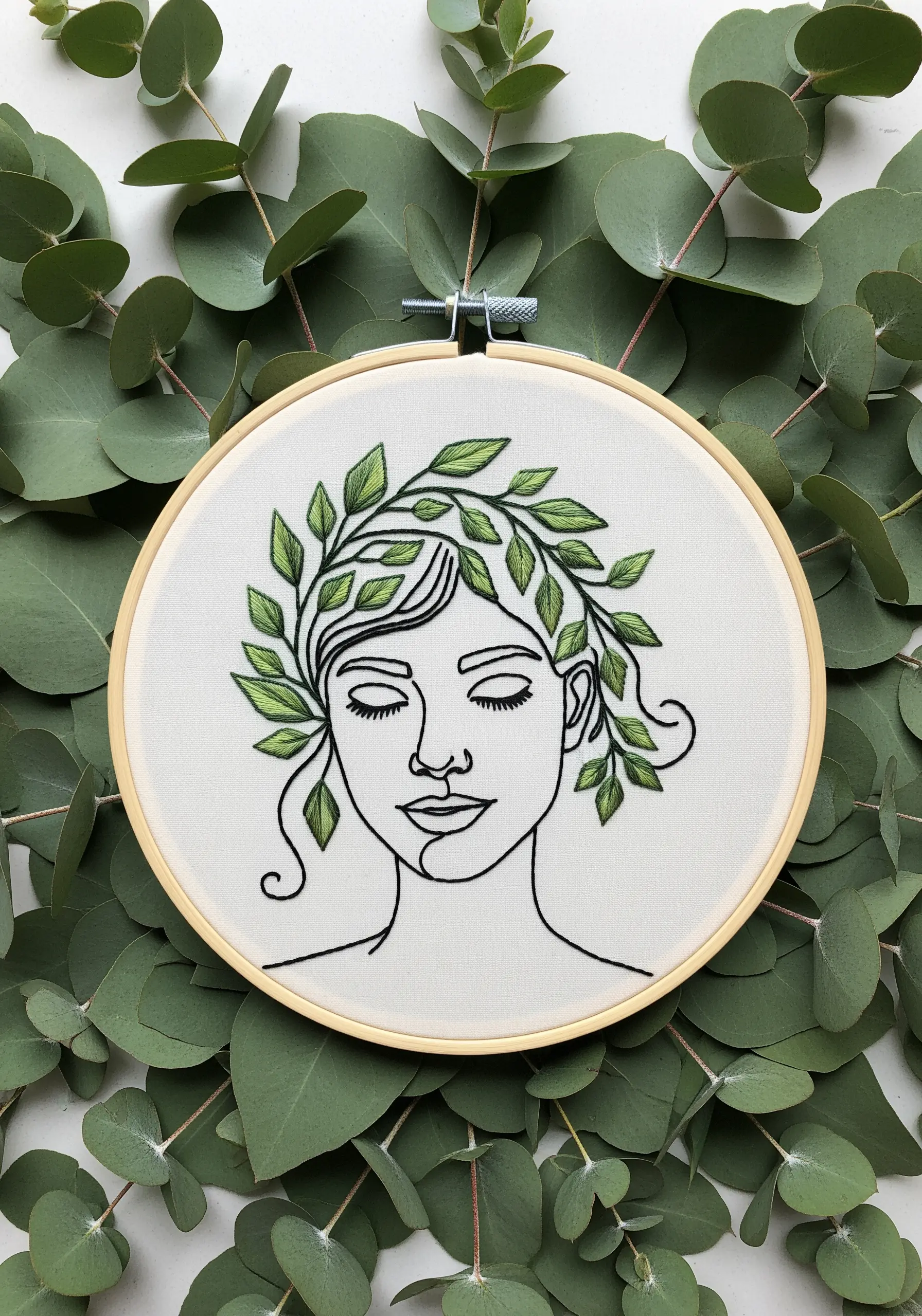 A minimal line art face crowned with satin-stitched green leaves, laid over real eucalyptus.