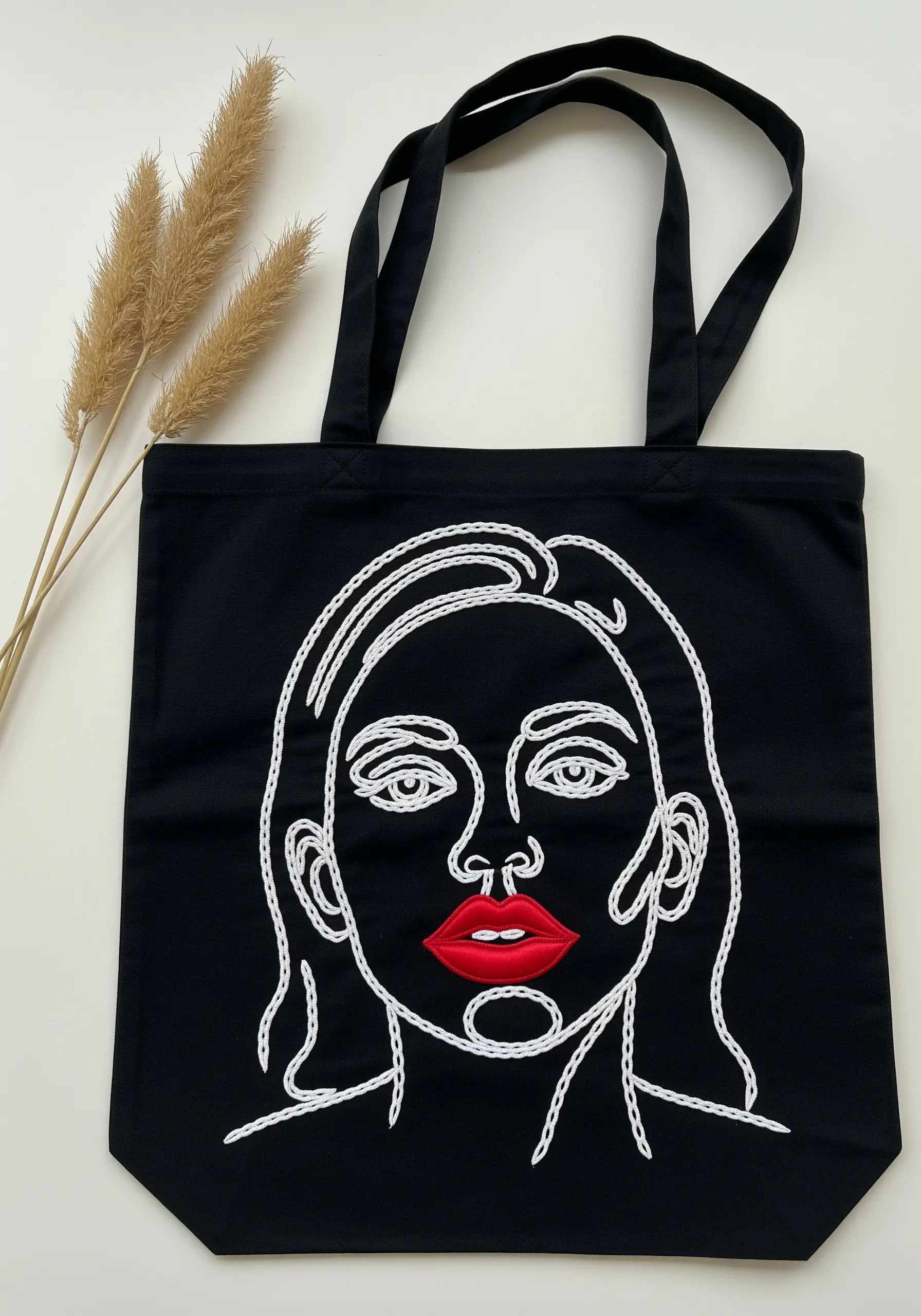 A white line art portrait on a black tote bag with bold, red satin stitch lips.