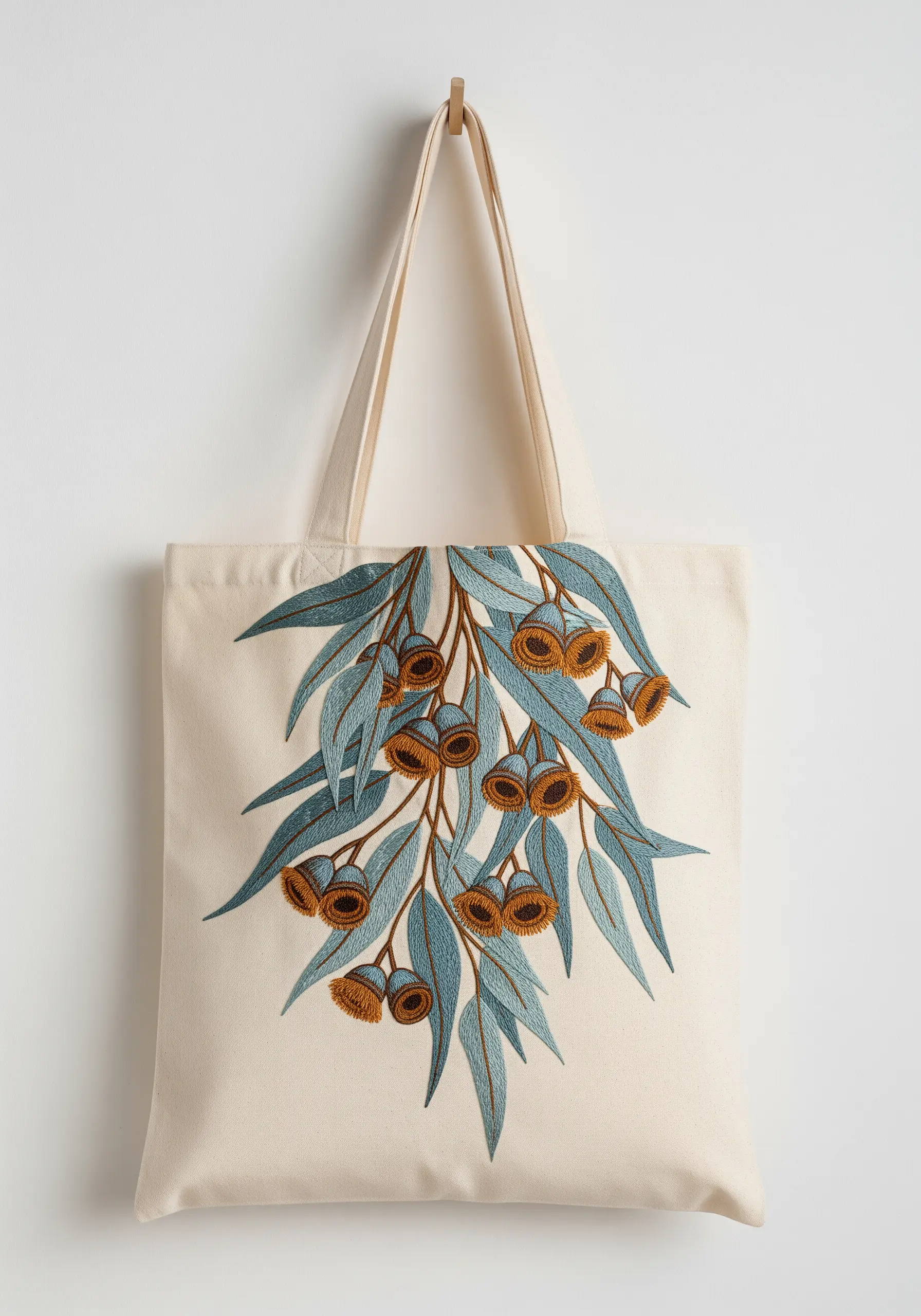 A cascading branch of embroidered eucalyptus leaves and gum nuts on a cream canvas tote bag.