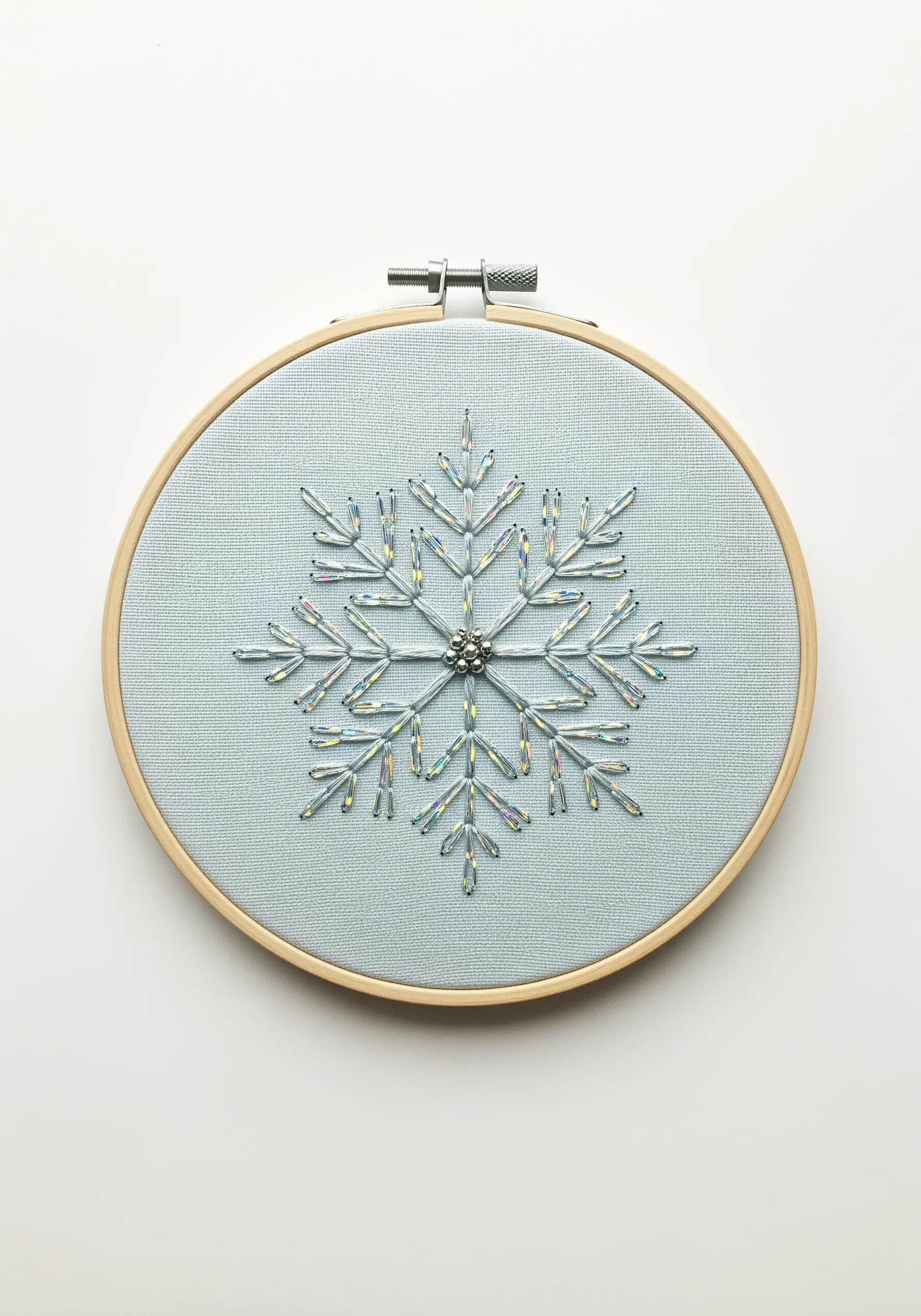 A snowflake motif created with iridescent bugle beads on a light blue fabric hoop.