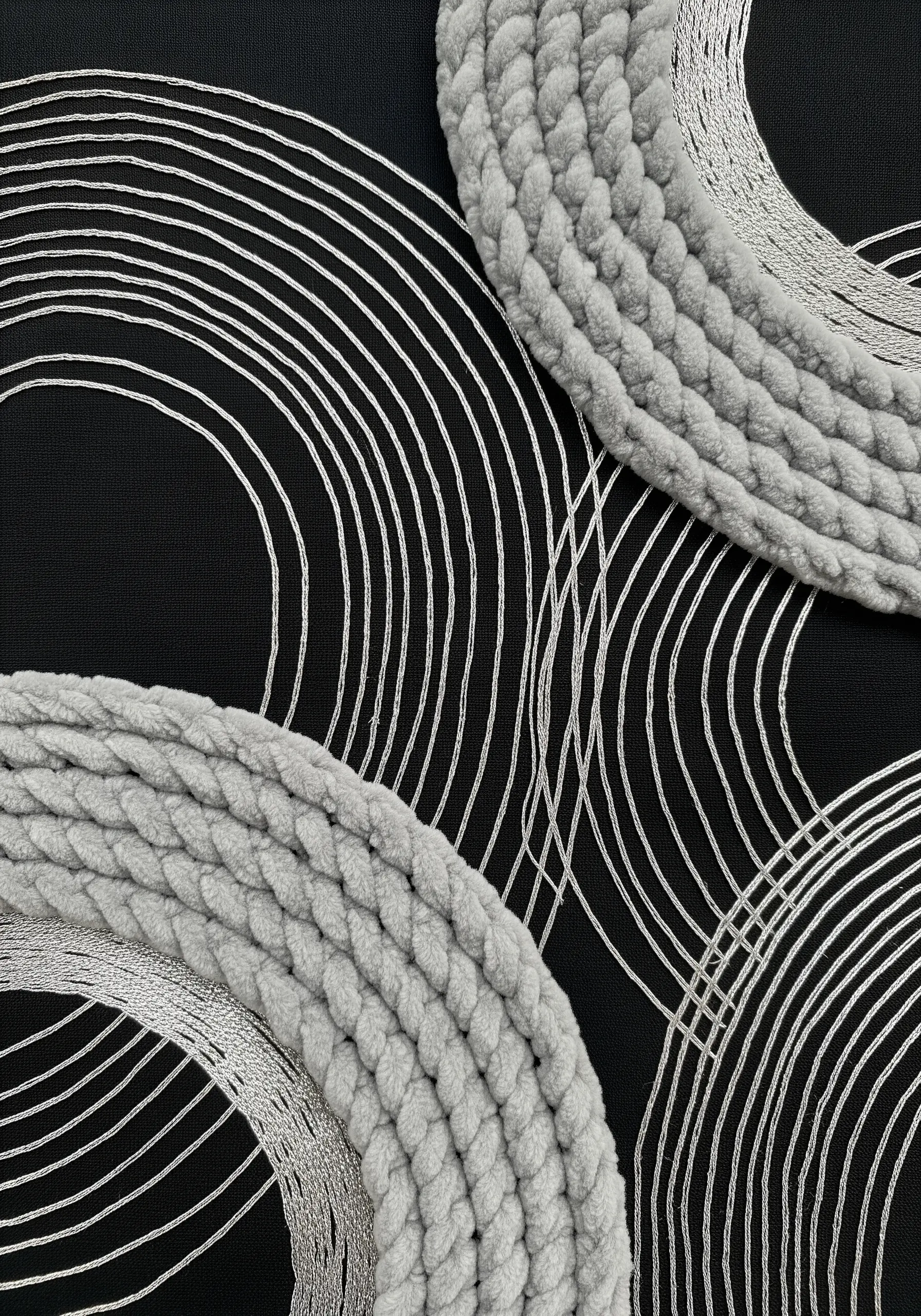 Close-up of thick grey braided cord next to fine, curving silver metallic threads on black fabric.
