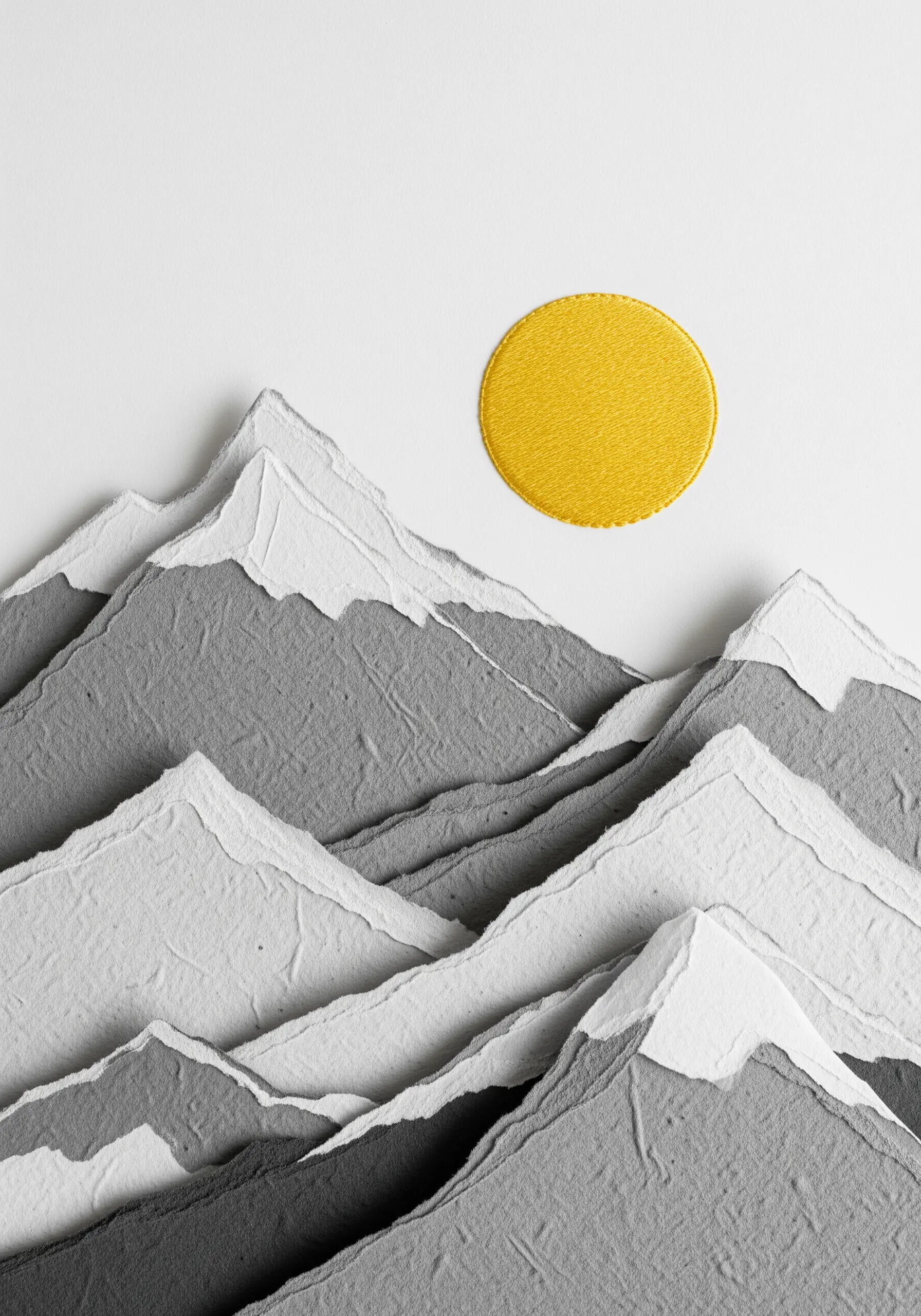 A mountain landscape made from torn grey and white paper, with a perfectly smooth yellow satin-stitched sun.