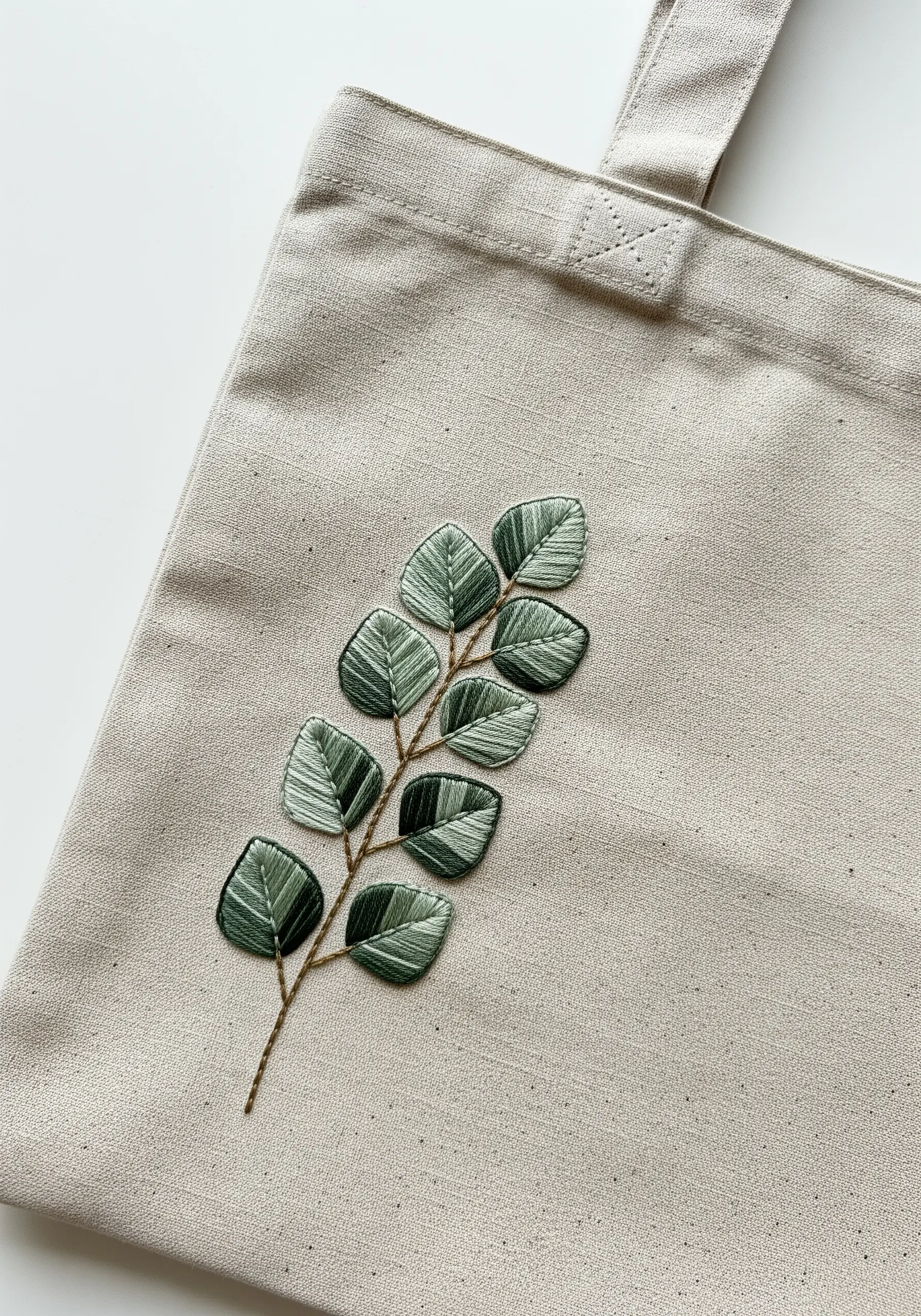 A sprig of leaves with green variegated thread on a natural linen tote bag.