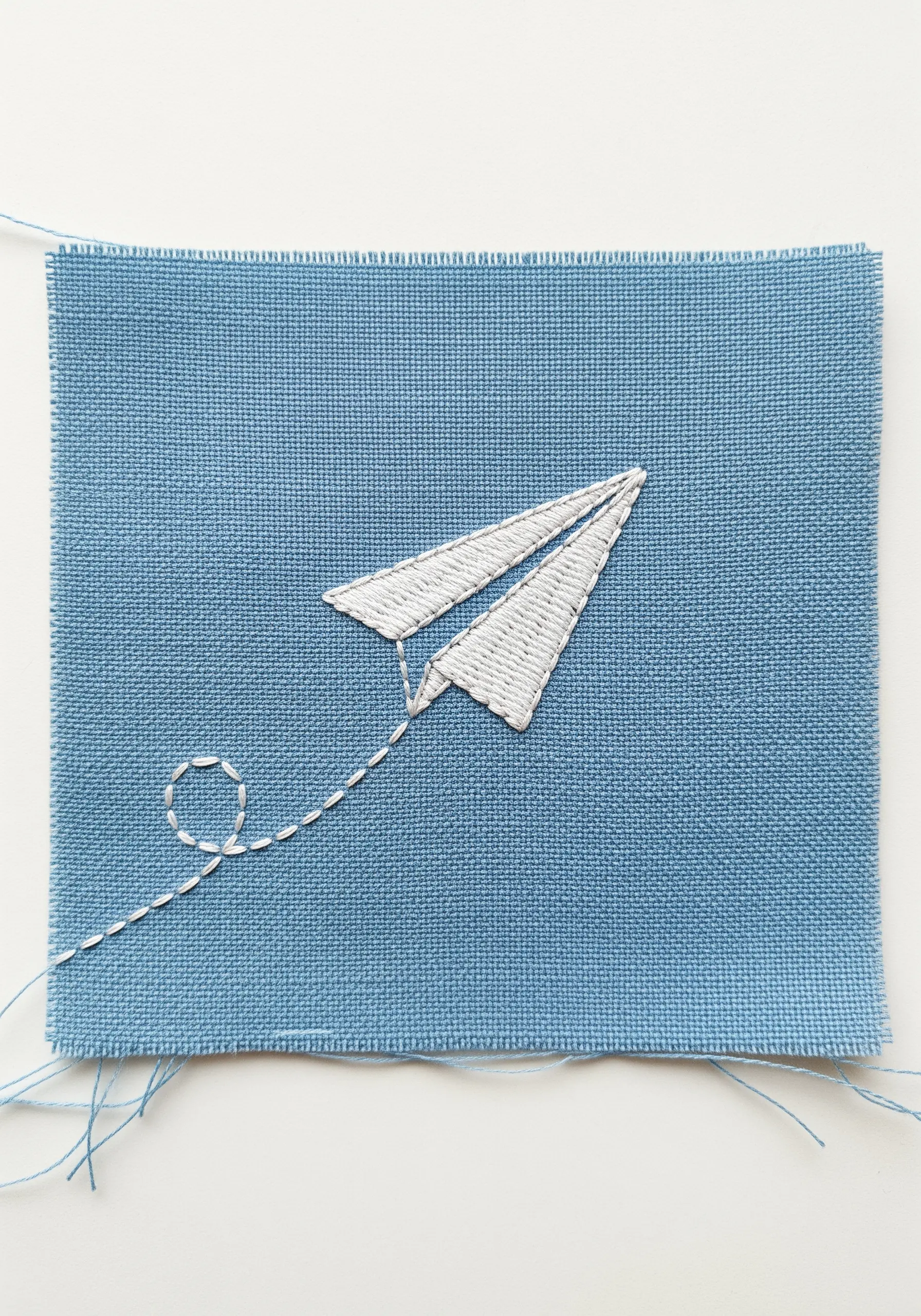 An embroidered paper airplane with a dashed line trail stitched on blue fabric.