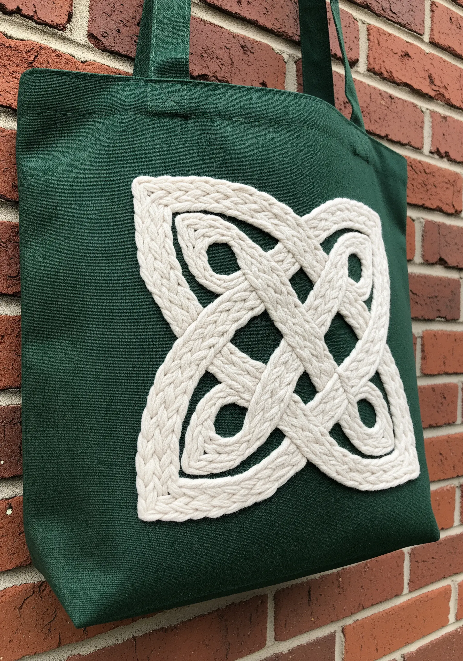 A large Celtic knot design made from thick, couched white cord on a green tote bag.