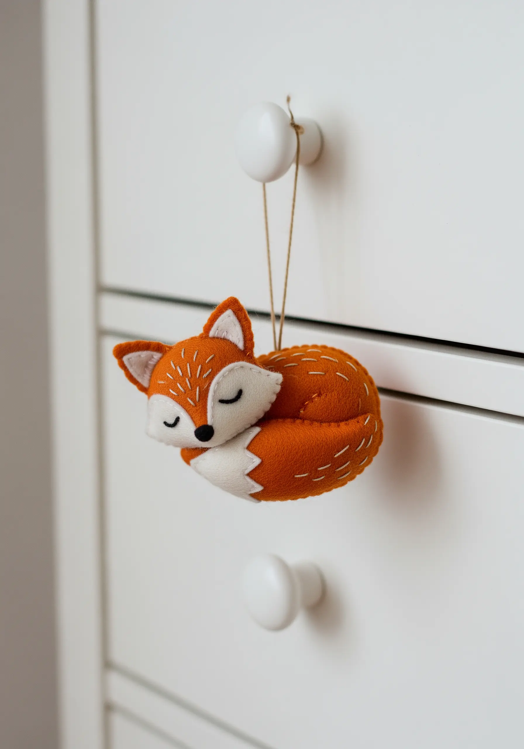 A small, three-dimensional felt fox ornament with white stitching, hanging from a drawer knob.