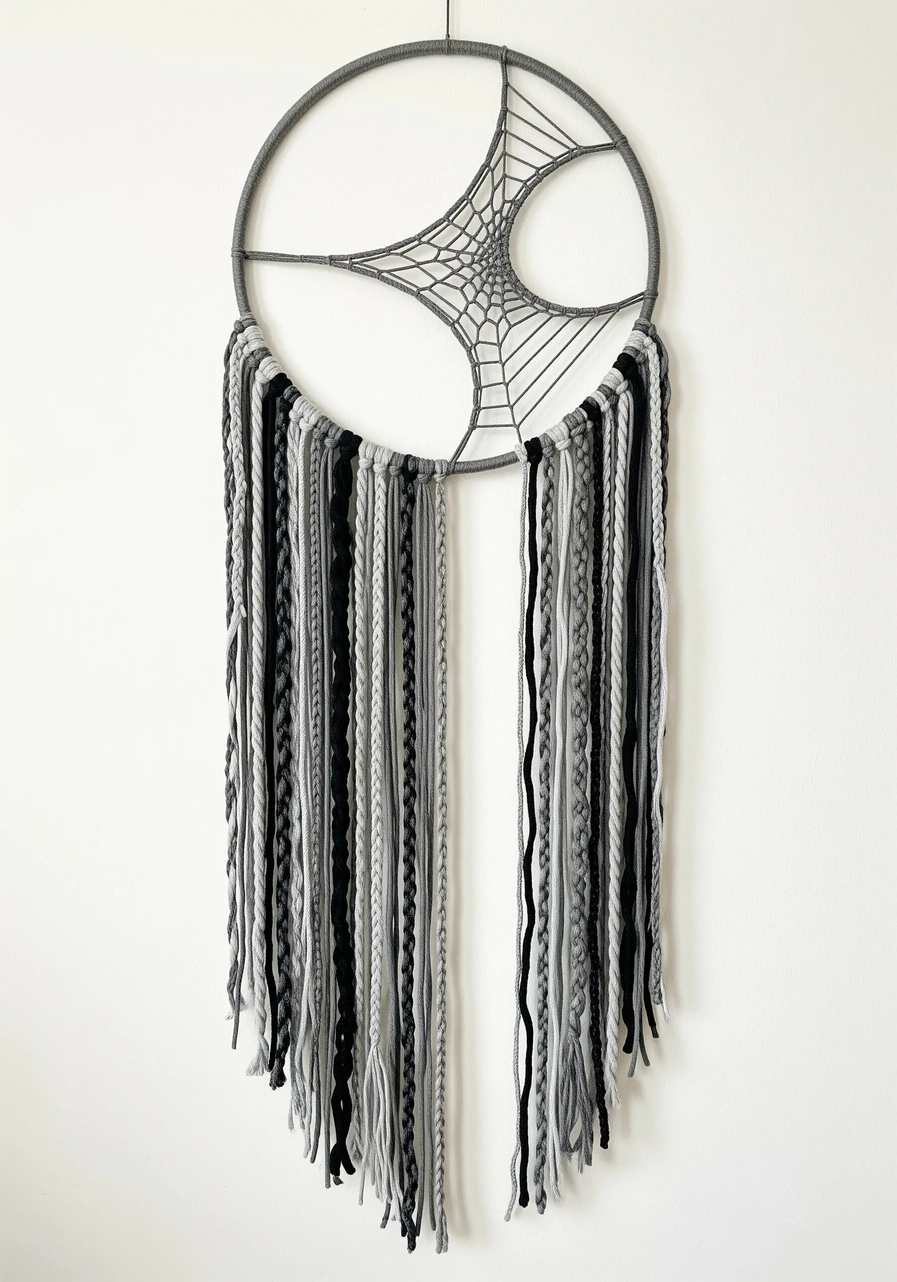 A modern dreamcatcher in shades of grey and black with an asymmetric web and braided tassels.