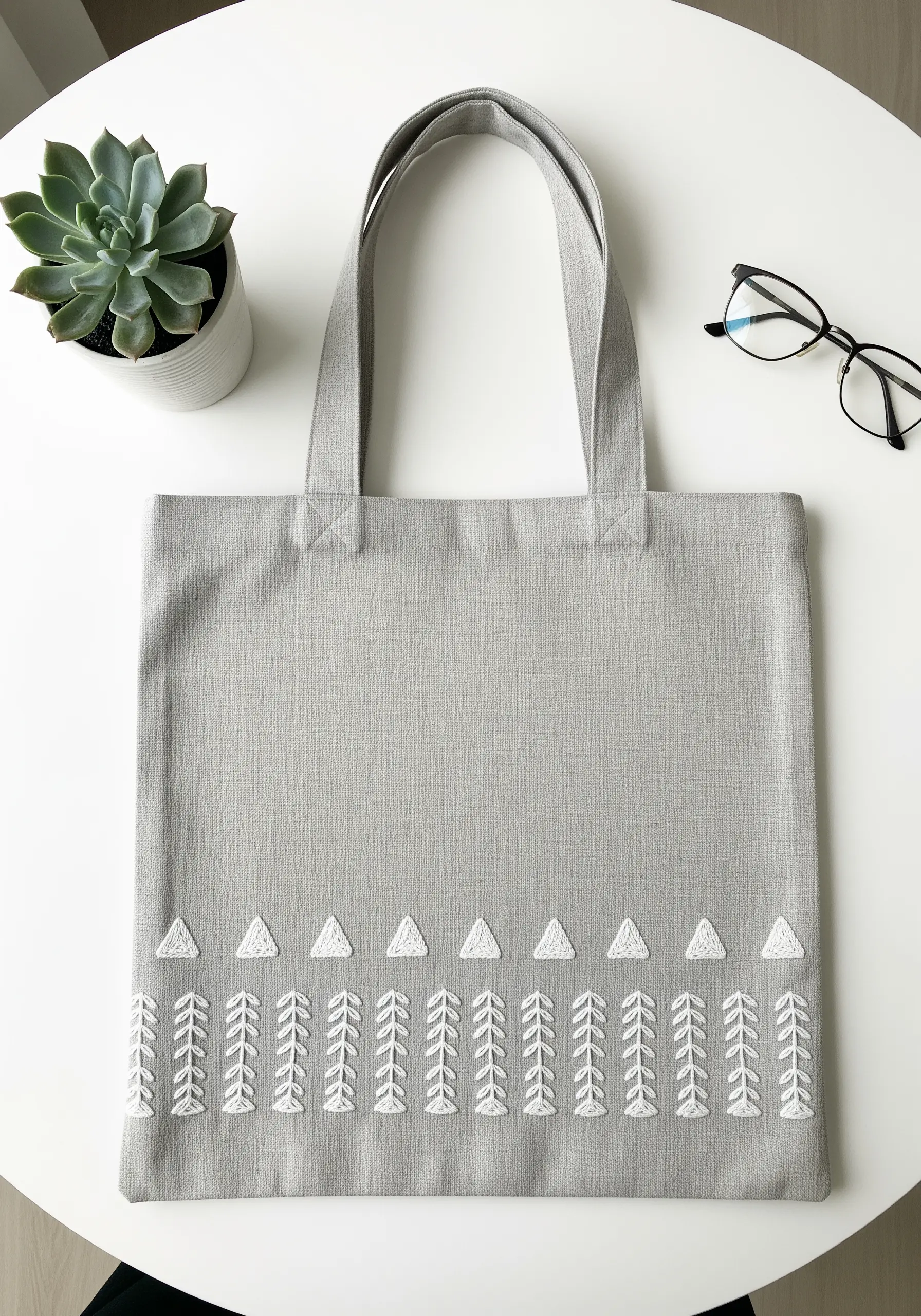 Minimalist white embroidered border with triangles and leaf motifs on a grey linen tote.