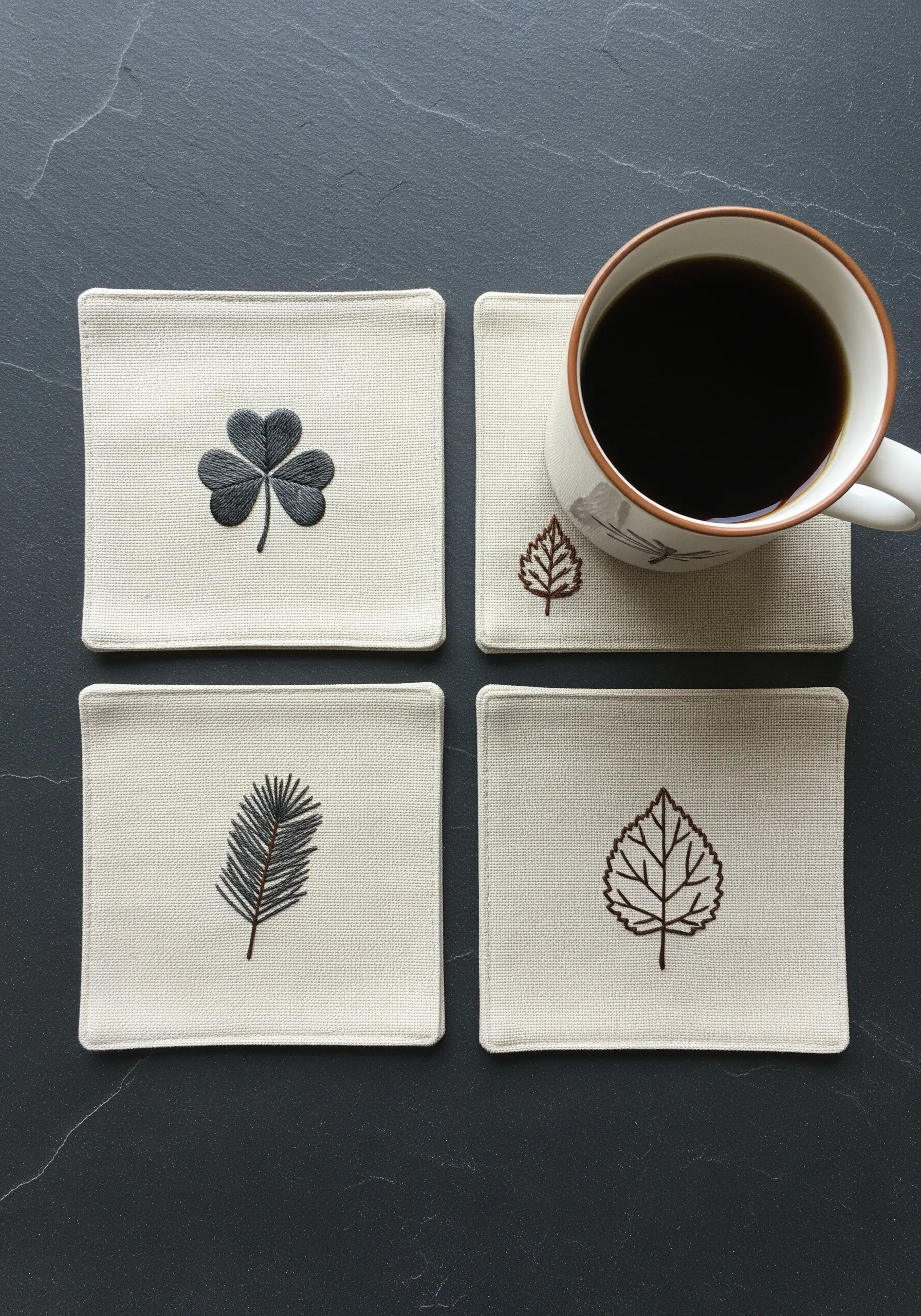A set of four linen coasters, each embroidered with a simple, dark leaf motif.