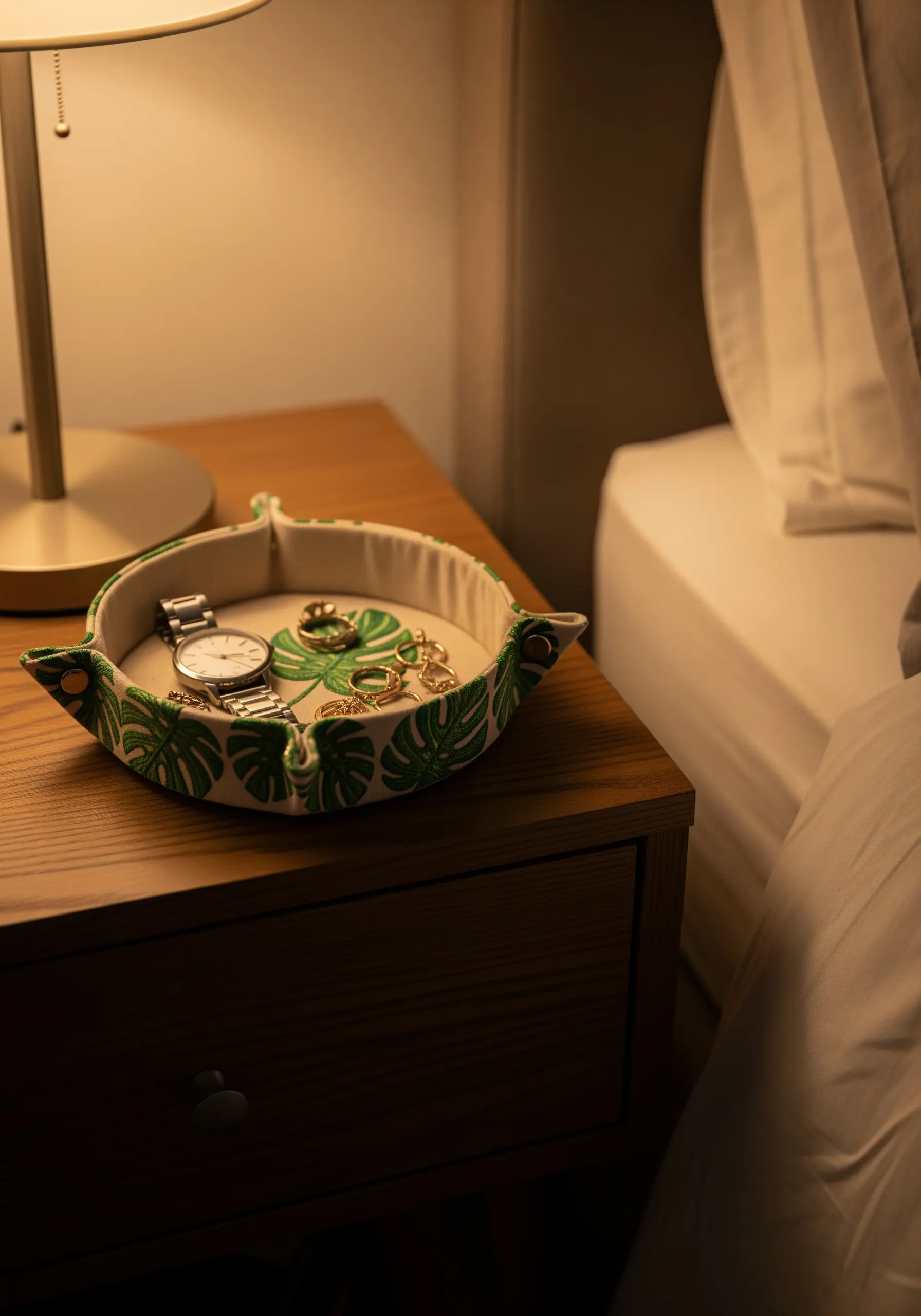 A fabric catch-all tray with embroidered monstera leaves holding jewelry on a nightstand.