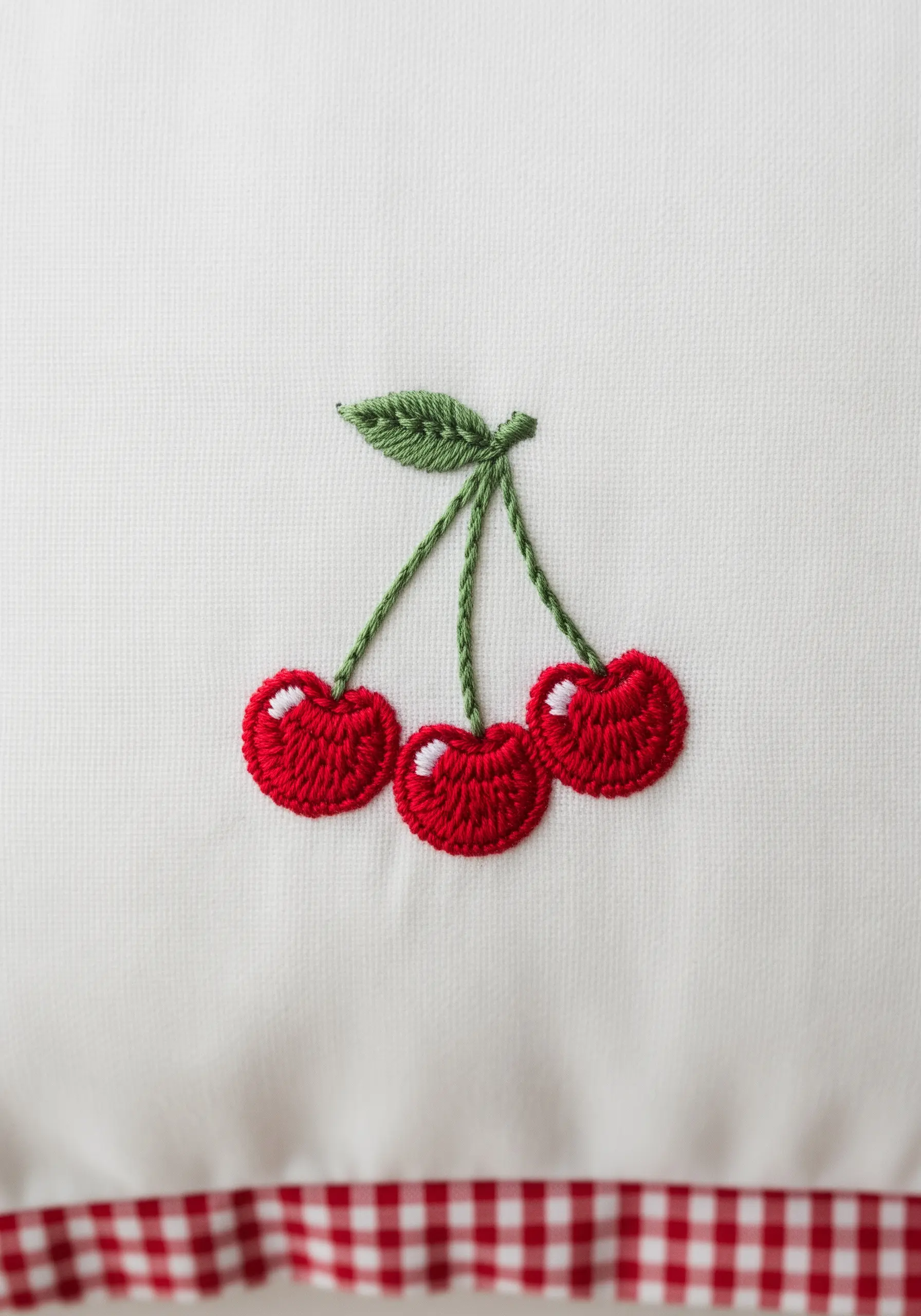 Three bright red cherries with a green leaf and stems, embroidered on a white and gingham pillowcase.