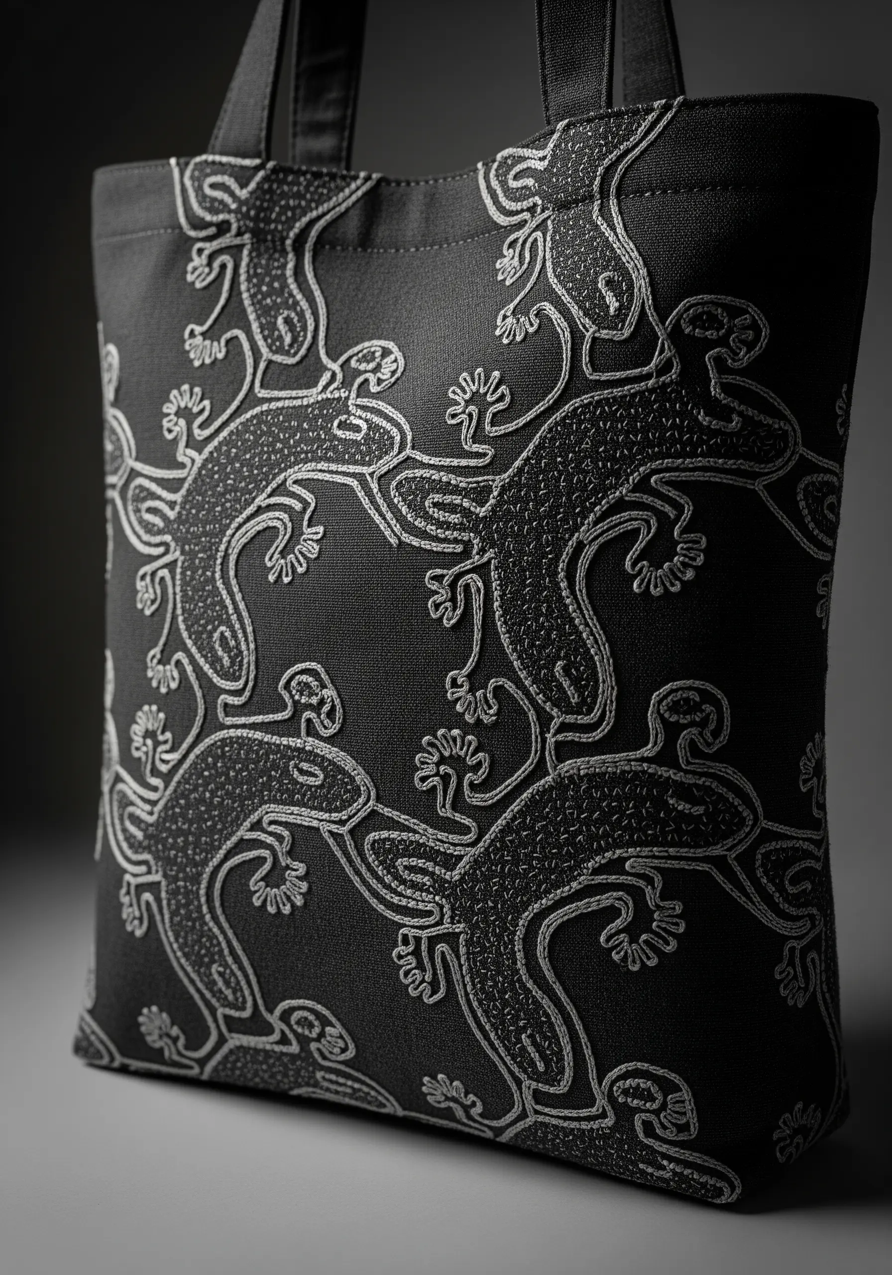 A tessellated pattern of interlocking lizards embroidered in white on a black canvas tote.