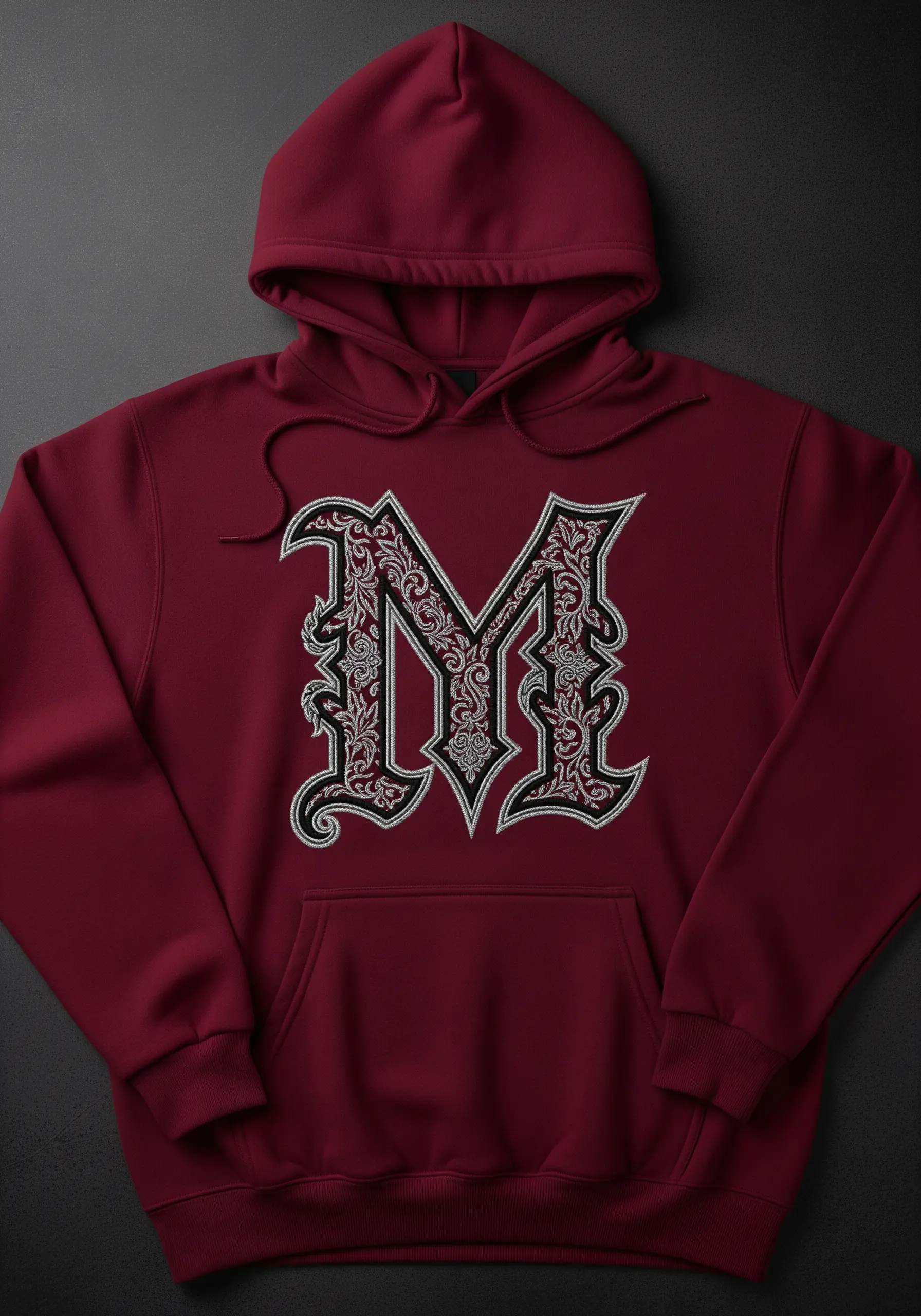 A large, ornate gothic letter 'M' embroidered in black and white on a burgundy hoodie.