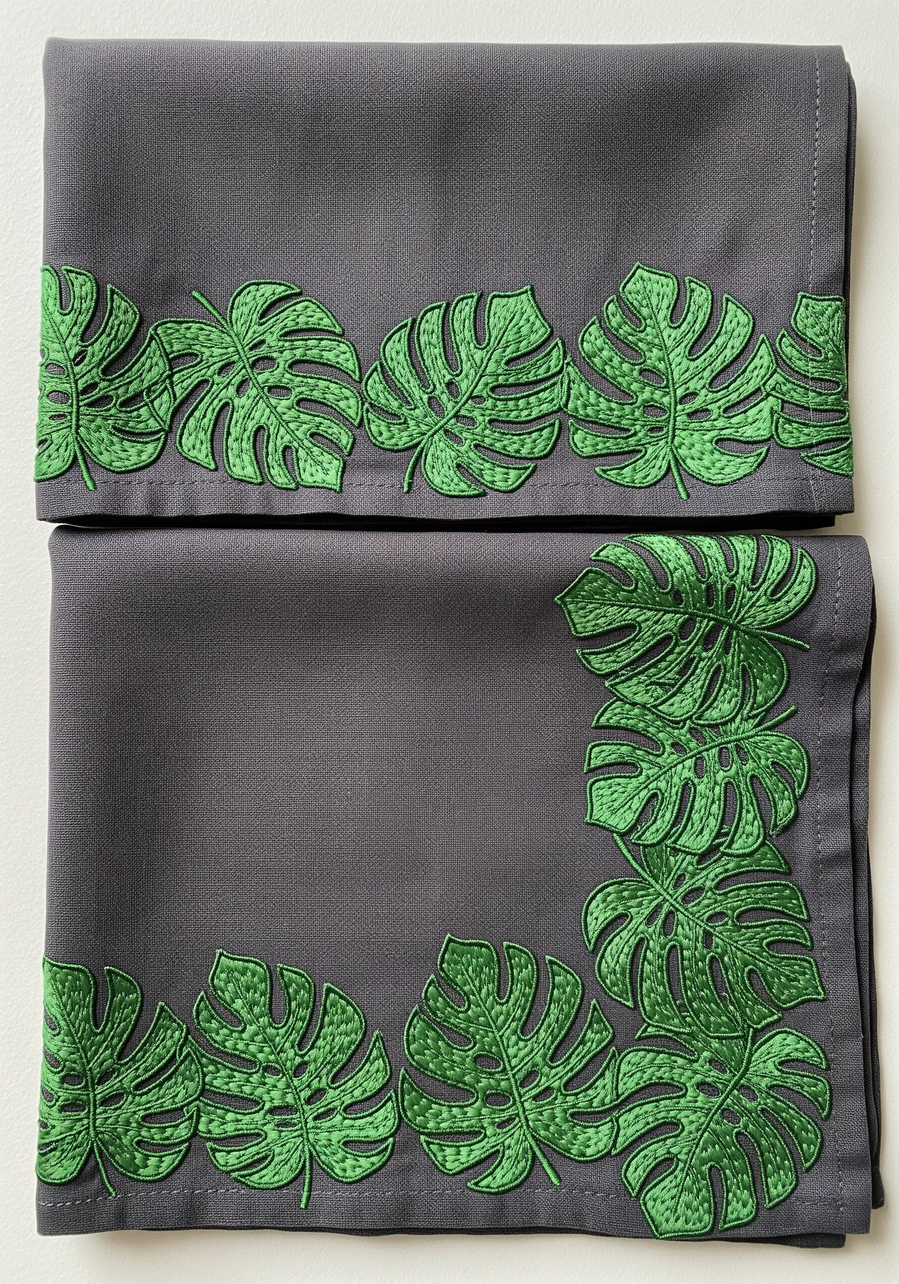 A border of bright green, graphic monstera leaves embroidered on a charcoal grey napkin.