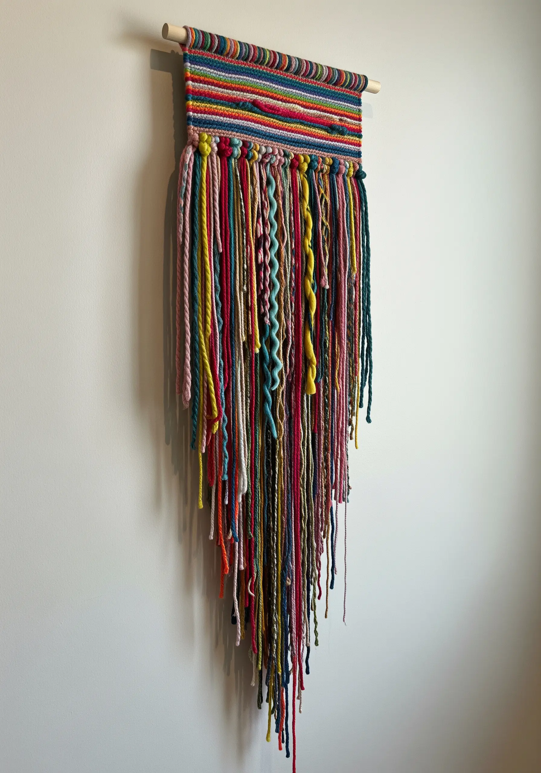 A colorful yarn wall hanging with a woven top and a long, cascading fringe of multi-colored yarns.
