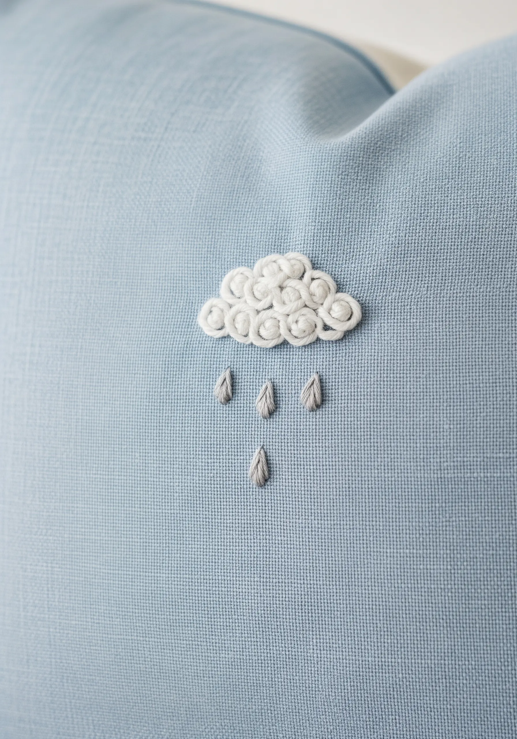A fluffy white cloud made with textured stitches, with several gray raindrops falling from it.