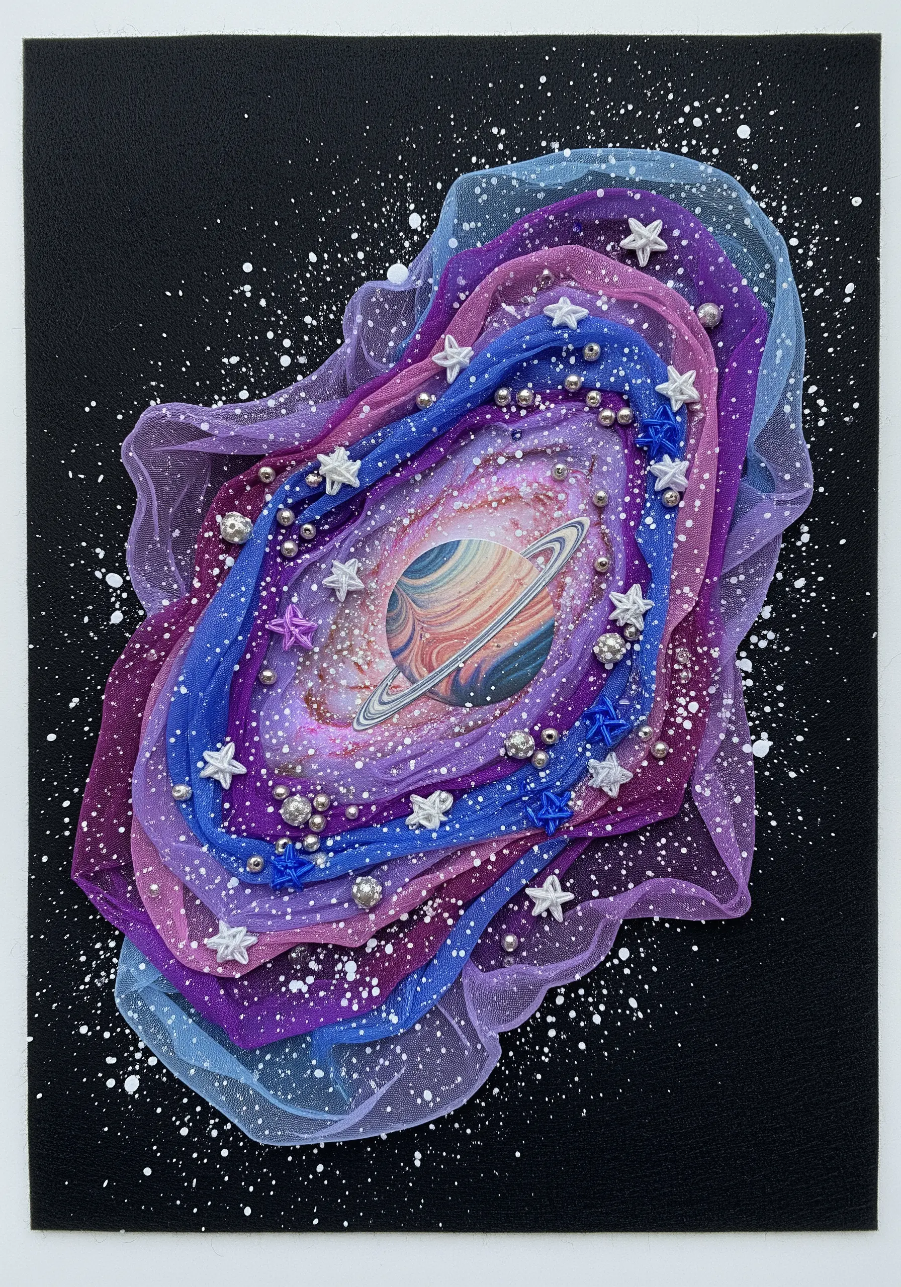 A layered tulle galaxy collage with a planet at the center, embellished with beads and stars.