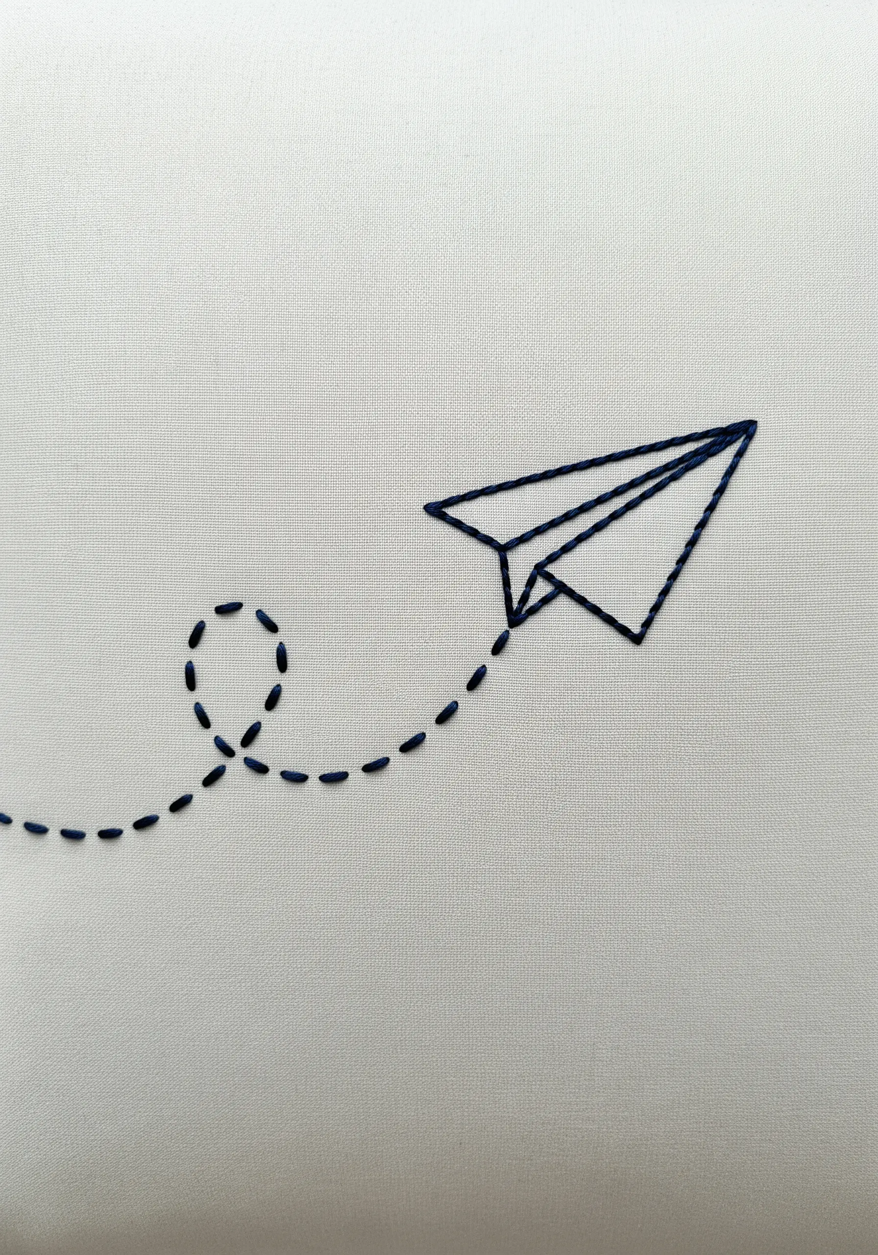 A paper airplane outlined in dark blue thread with a dotted flight path behind it.