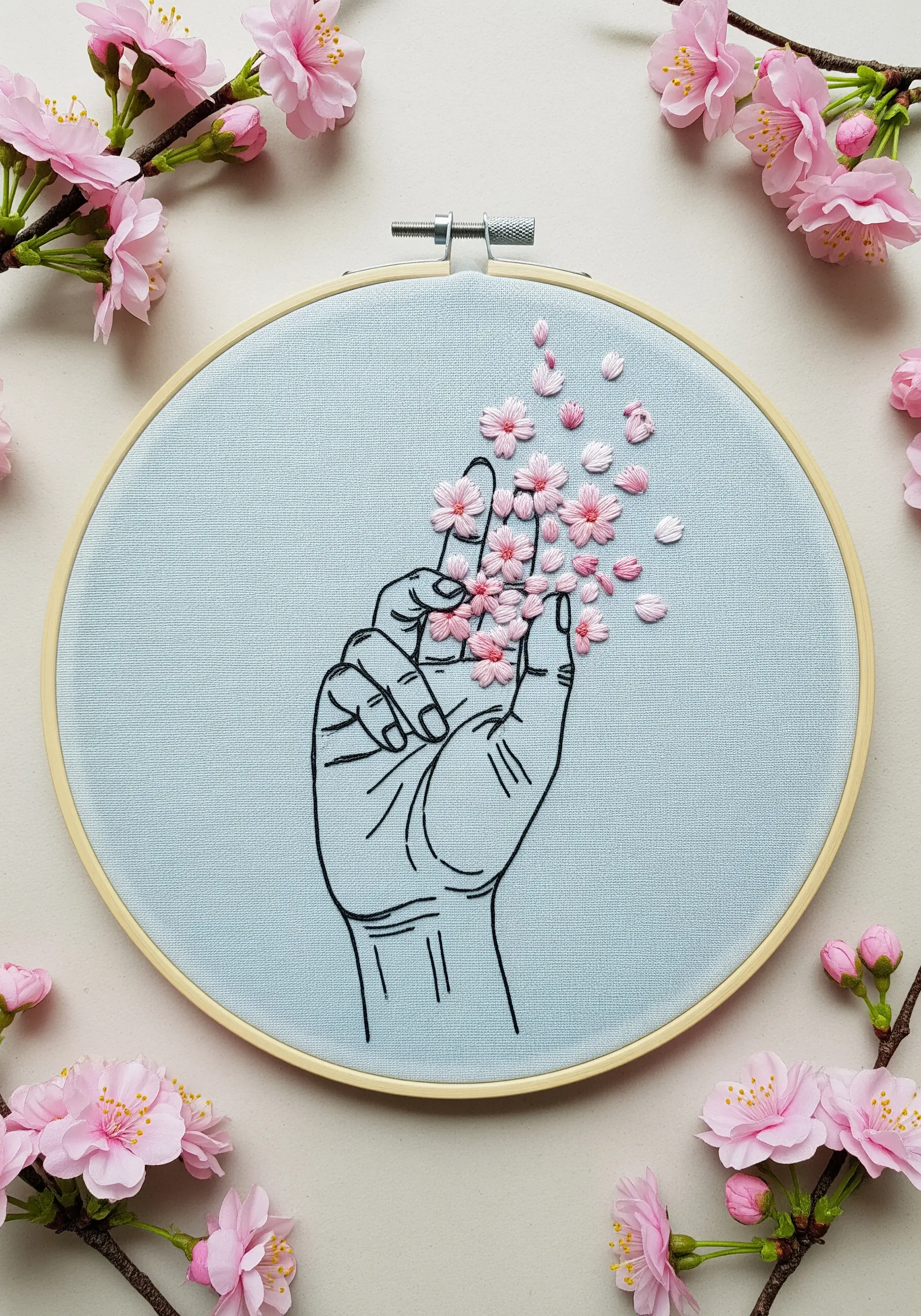 Line art hand with pink cherry blossom petals floating upwards on light blue fabric.