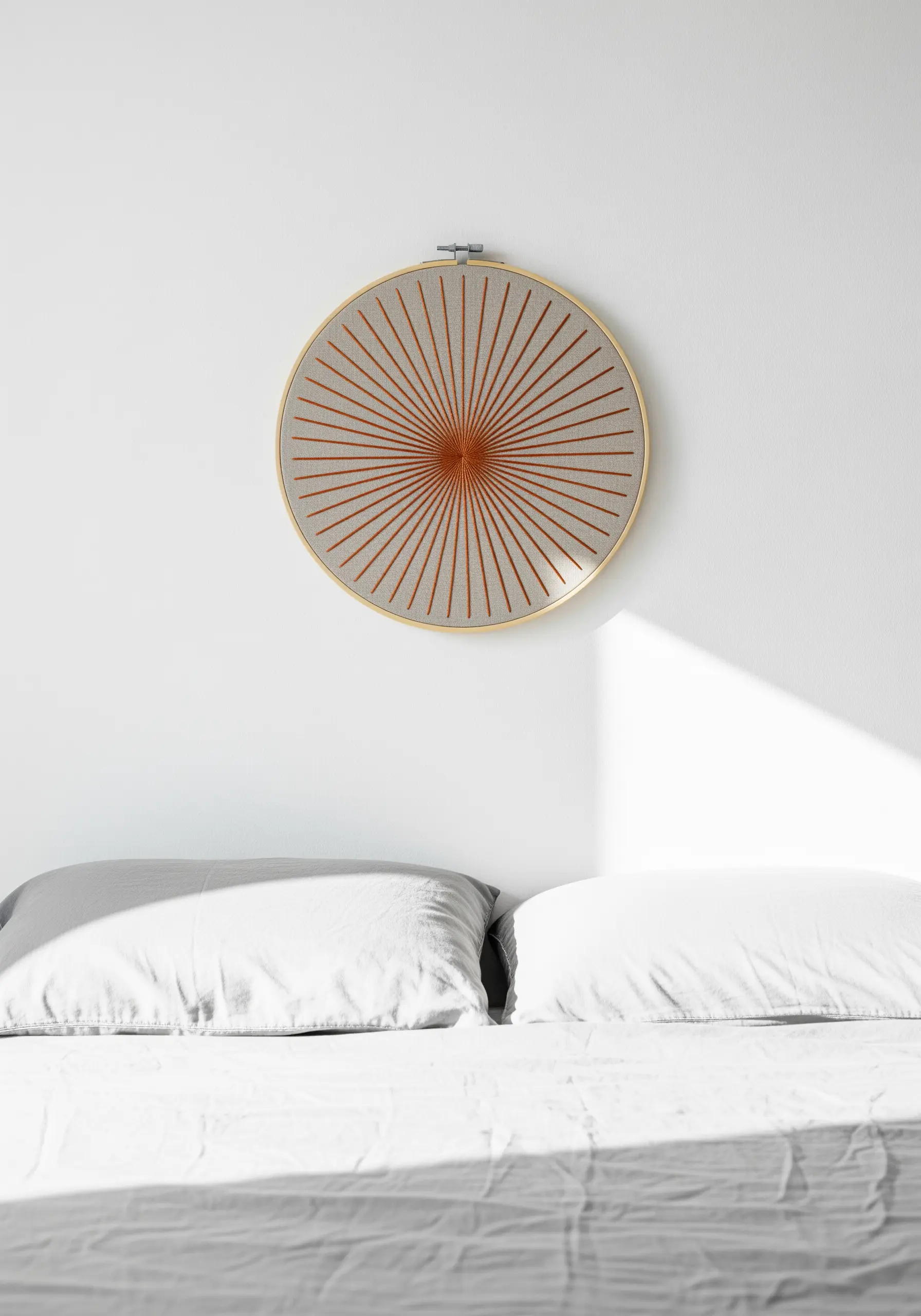 A geometric sunburst embroidered with terracotta thread in a hoop over a bed.