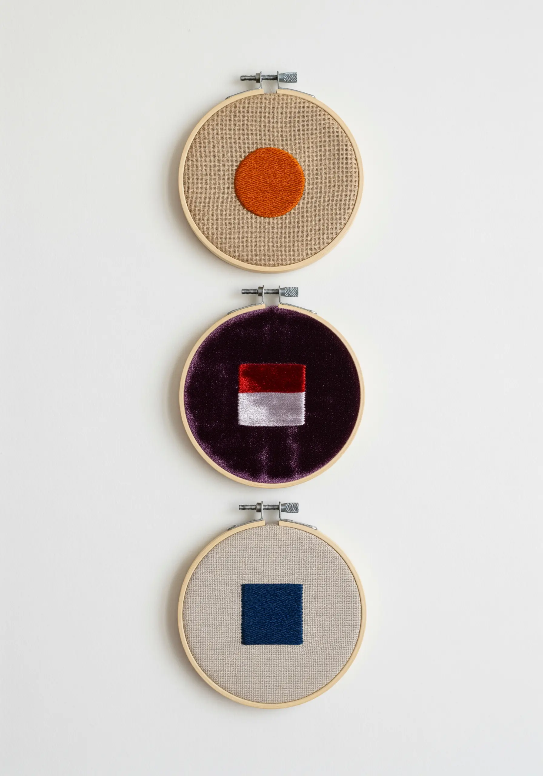 A triptych of three embroidery hoops with simple geometric shapes on different textured fabrics.