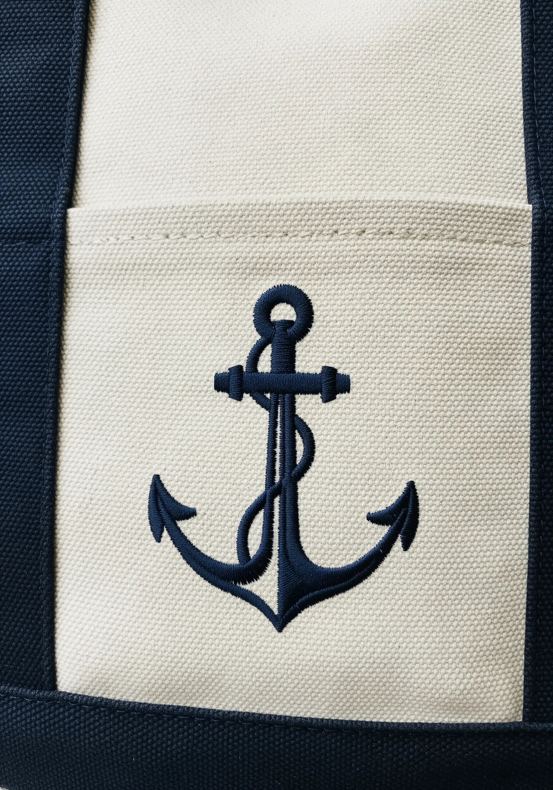 A navy blue anchor embroidered with a raised satin stitch on the pocket of a canvas tote bag.