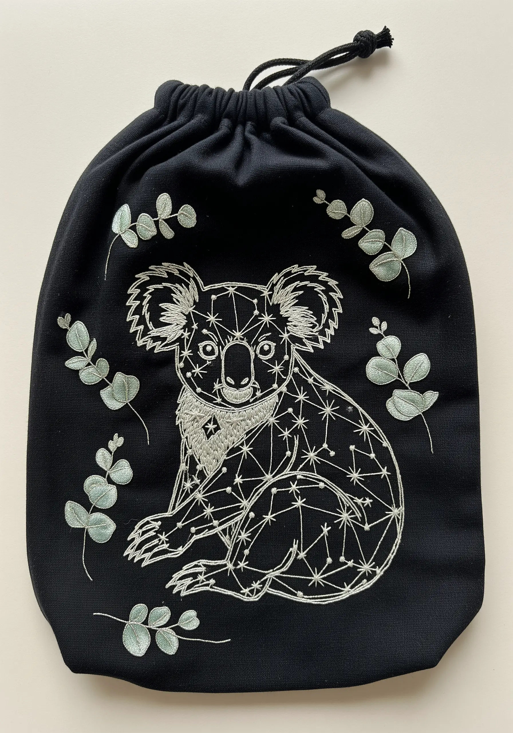 A geometric koala constellation embroidered in silver thread on a black drawstring bag.