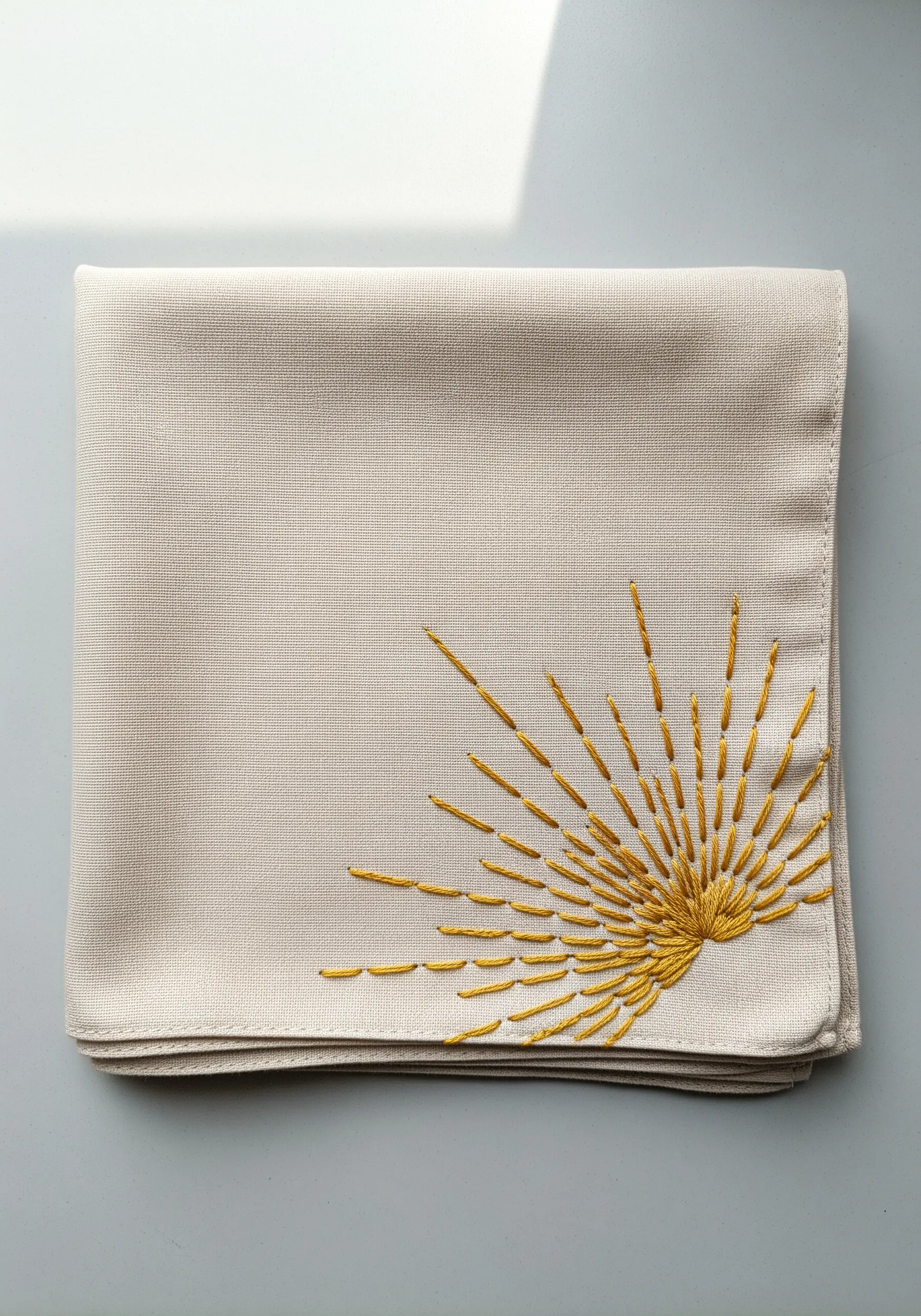 A golden sunburst motif with radiating straight stitches embroidered in the corner of a beige handkerchief.