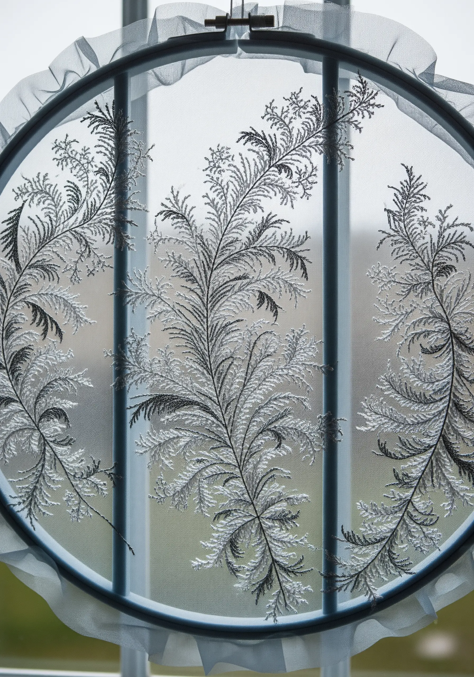 Delicate frost-like ferns embroidered in white and grey thread on sheer fabric in a hoop.
