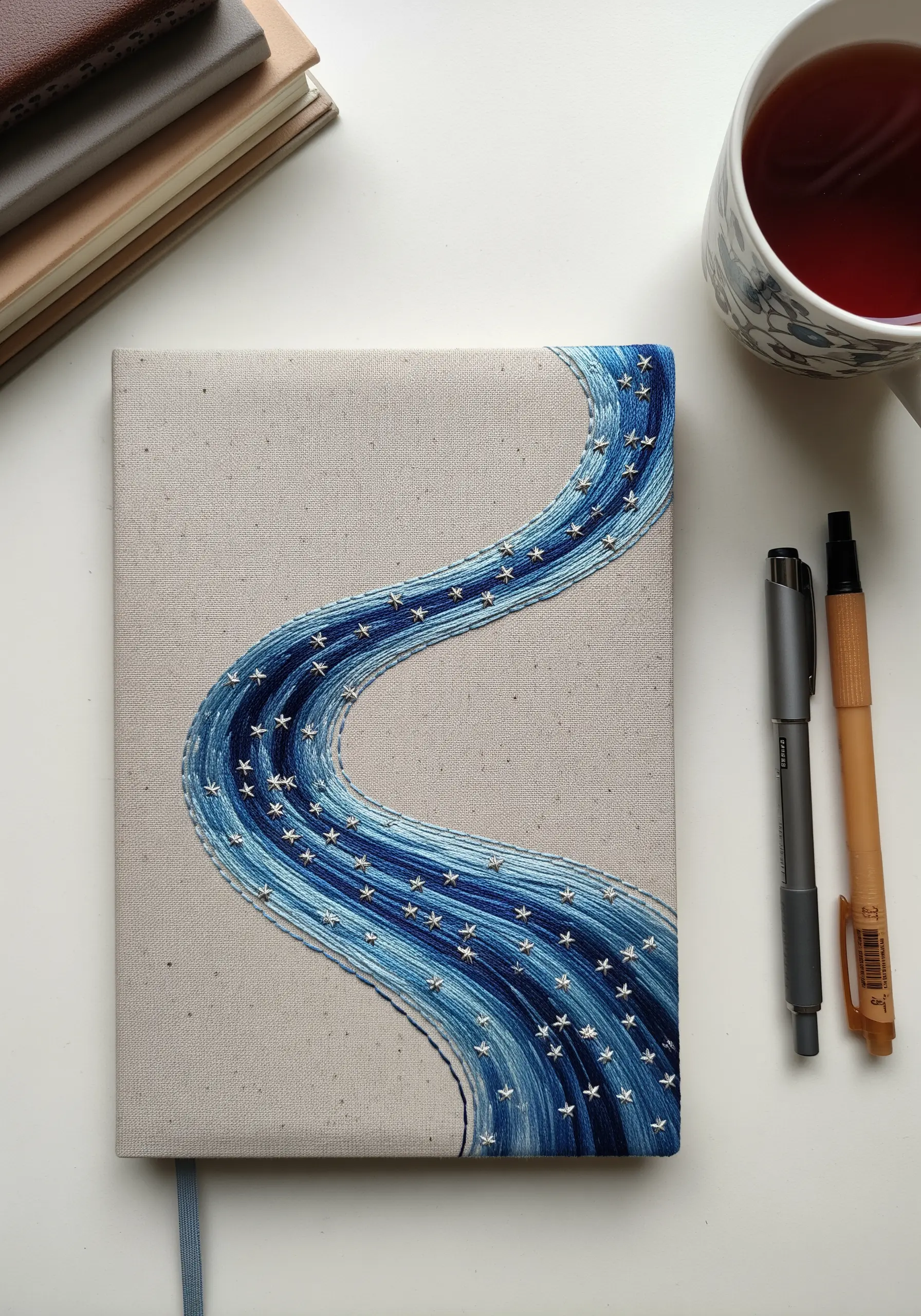 A fabric journal cover with an embroidered flowing river of blue gradient threads and white stars.